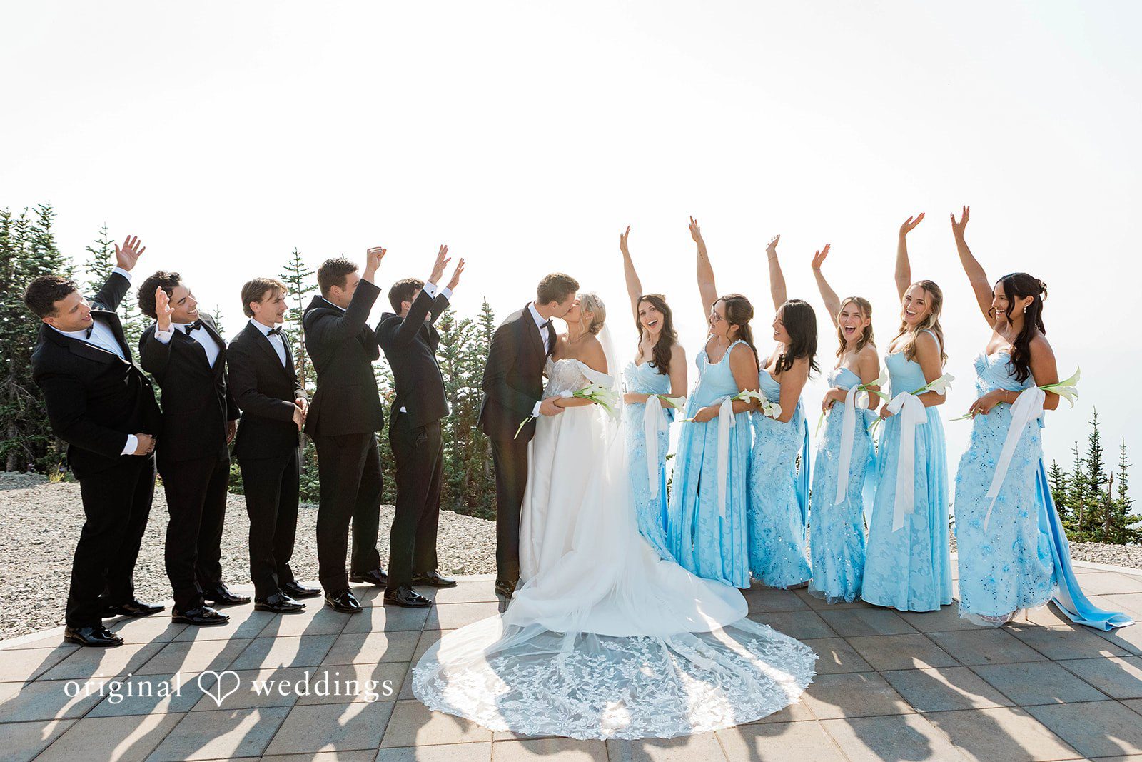 Bethany + Ryan A wedding party stands in a line outdoors cheering with their arms raised.
