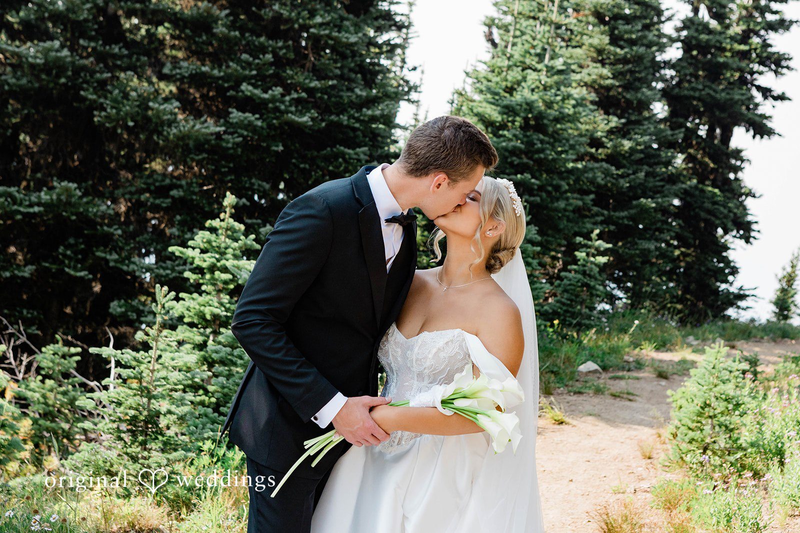 Bethany + Ryan A bride and groom share an intimate embrace and kiss in front of evergreen trees.