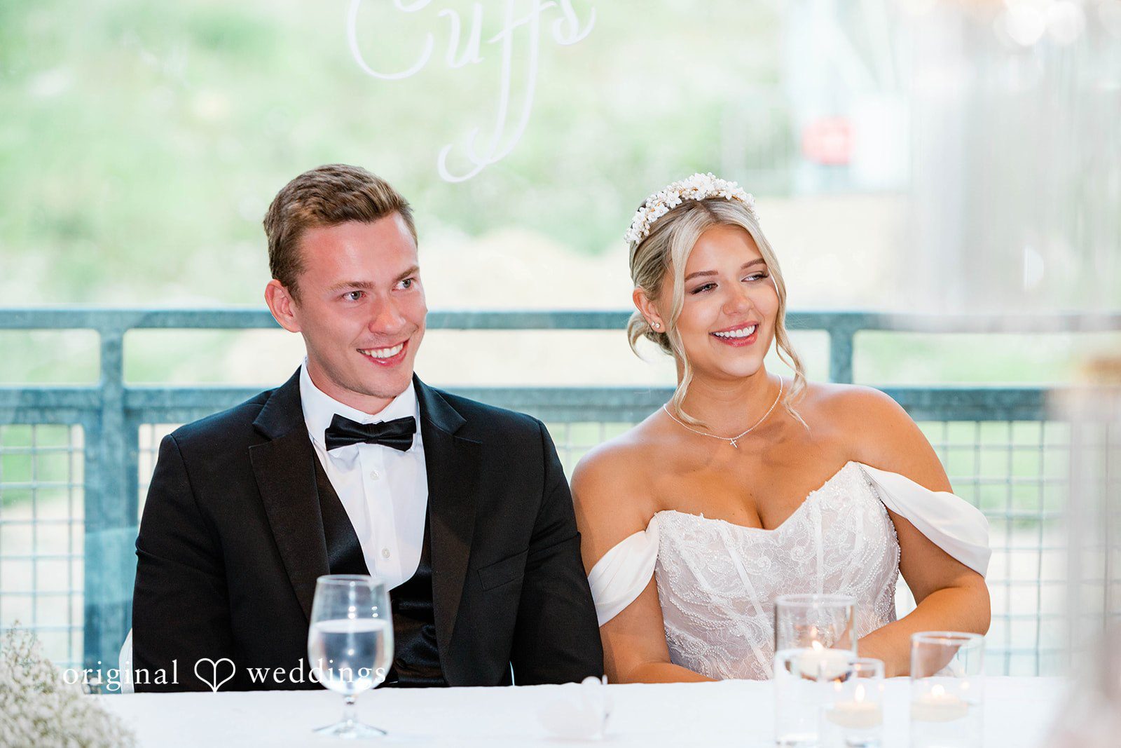 Bethany + Ryan A bride and groom sit at a decorated head table smiling during reception speeches.