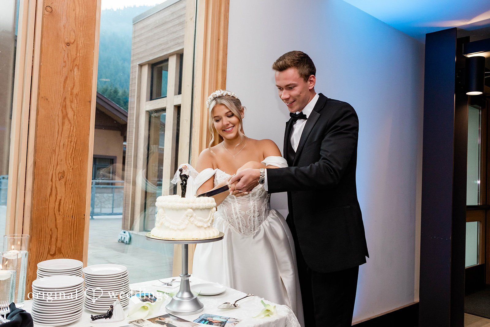 Bethany + Ryan A bride and groom stand together to cut their white tiered wedding cake.