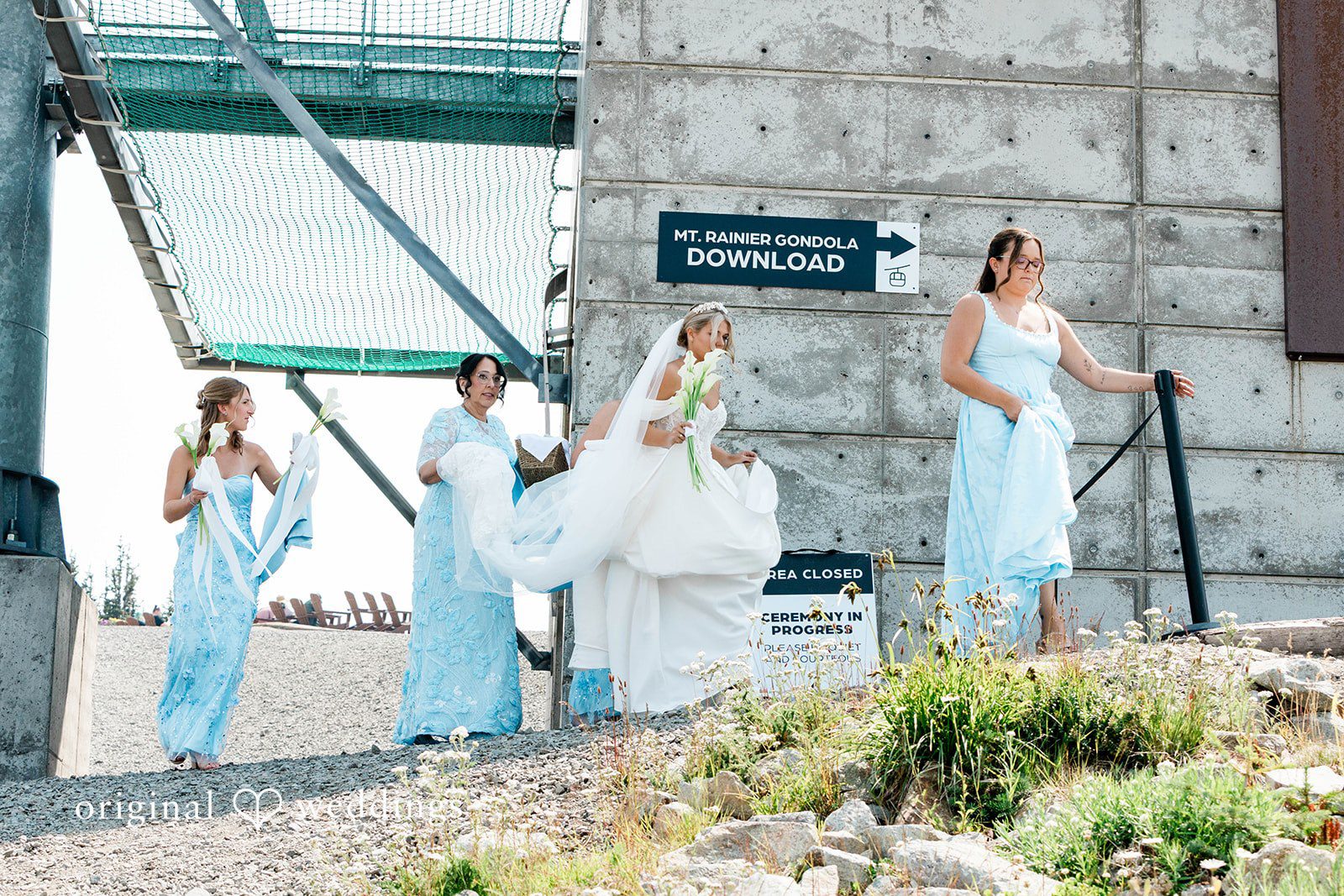 Bethany + Ryan A bride walks with her bridesmaids in light-blue dresses near a concrete structure.