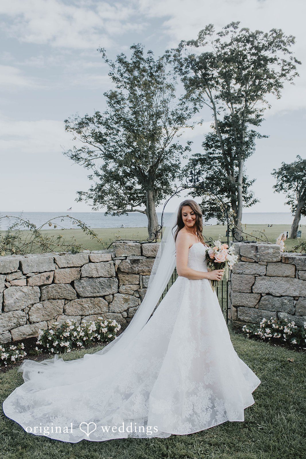 Alexa + Frank Bride holding her bouquet by the stone wall near the water at Eolia Mansion