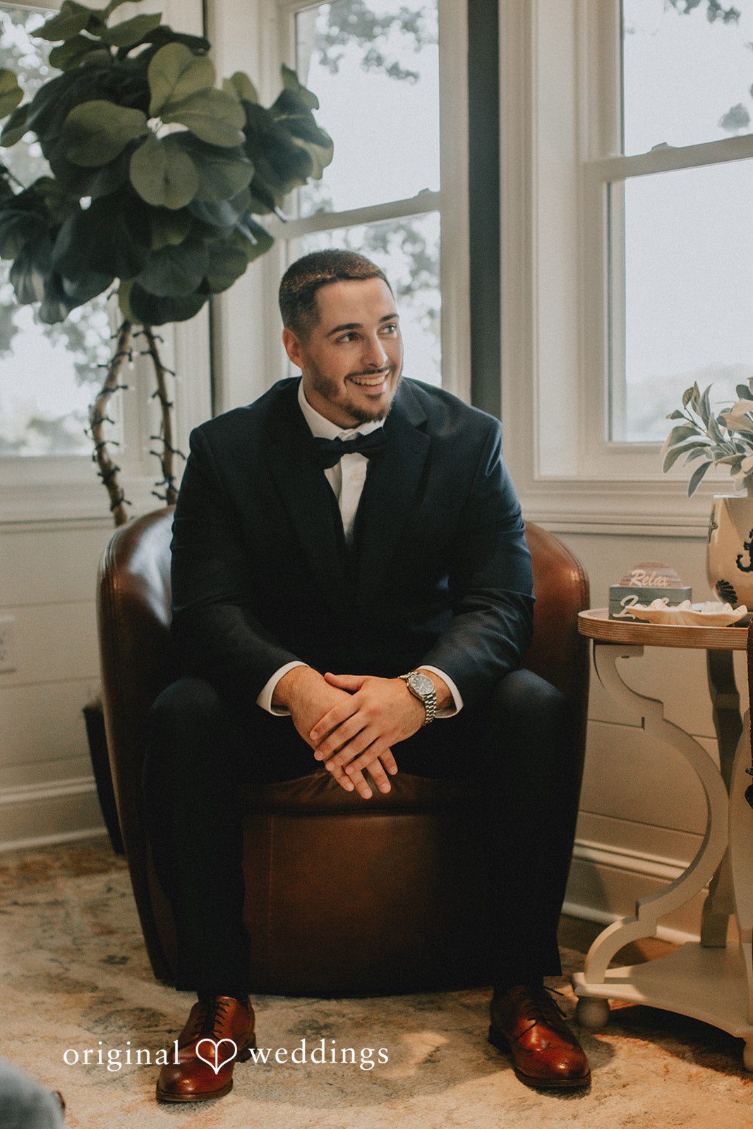 Sarah + Christopher A groom in a dark suit sitting alone on a chair in front of a window.