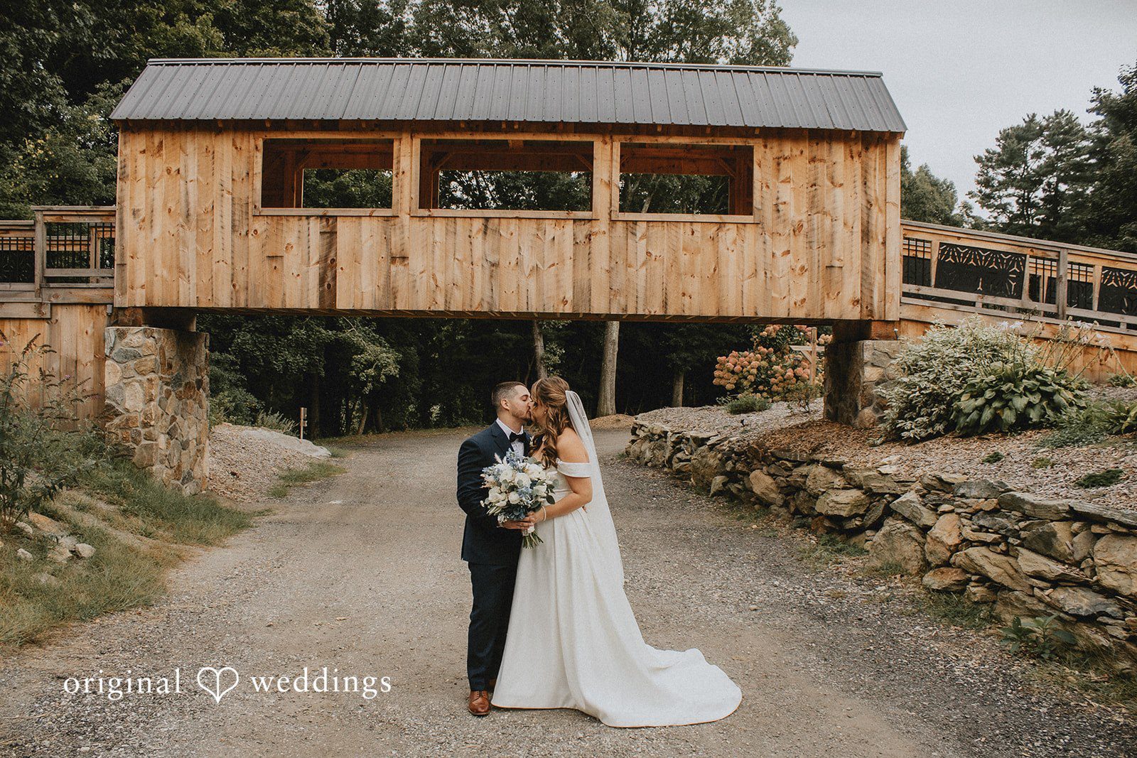 Sarah + Christopher A bride holding a bouquet and a groom in a dark suit share a kiss on a gravel path beneath a rustic wooden covered bridge structure.