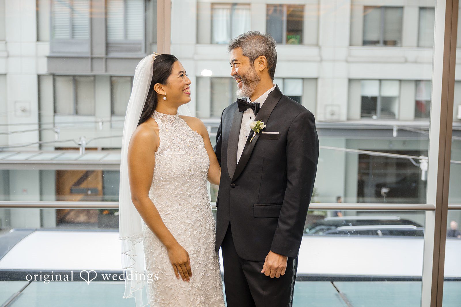 John + Michelle Bride in a sleeveless gown and groom in a dark suit standing together near a modern window.