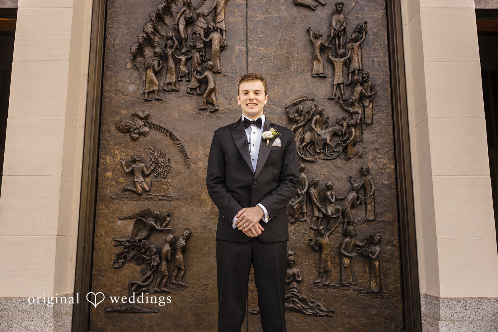 John + Michelle Groom dressed in a classic tuxedo standing before large ornate doors.