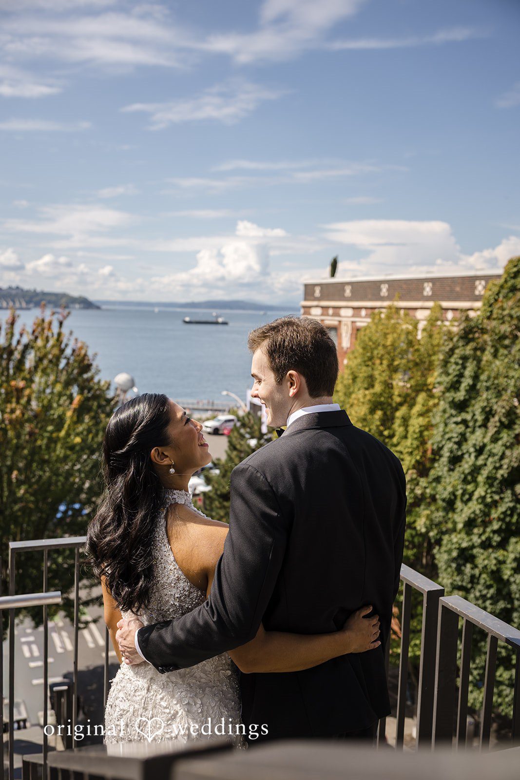 John + Michelle Bride and groom embracing on a balcony overlooking water and trees.