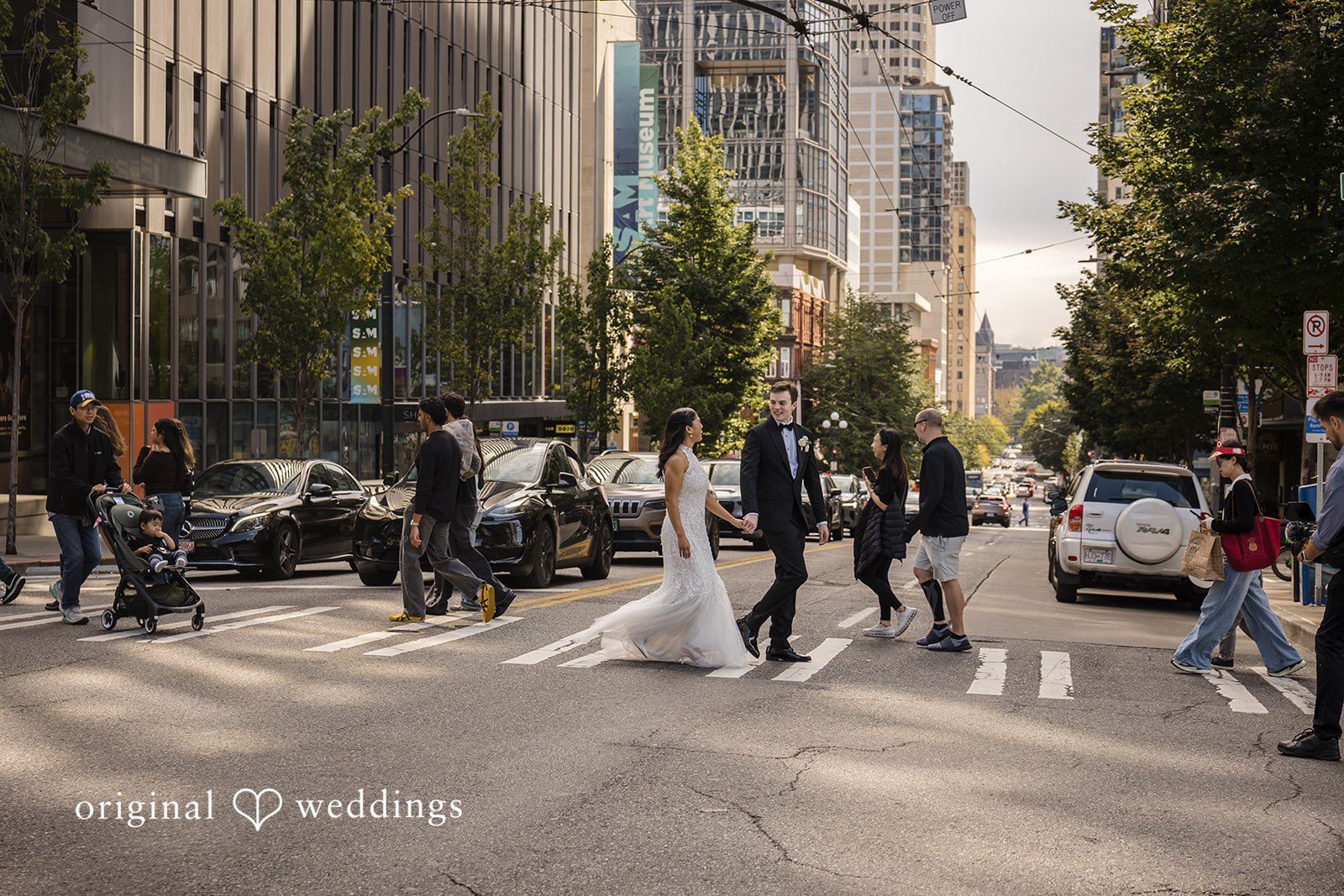 John + Michelle Bride and groom walking across a city street sidewalk with urban buildings in the background.