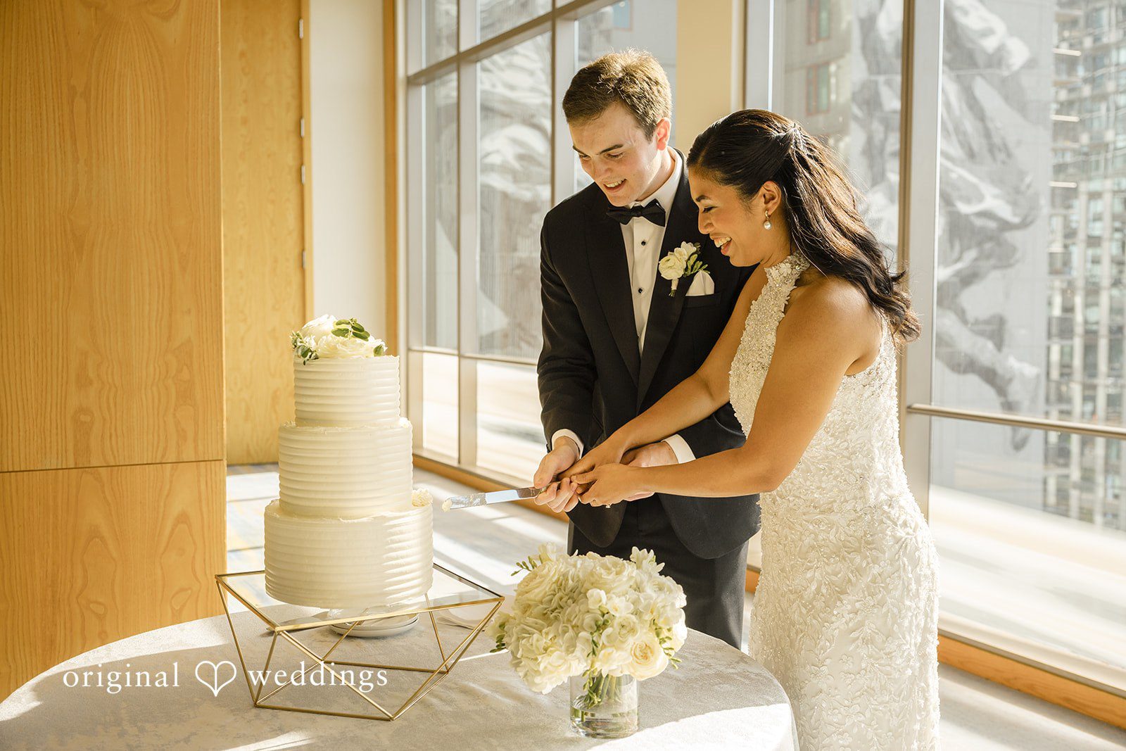 John + Michelle Bride and groom standing next to their tiered wedding cake near a large window.