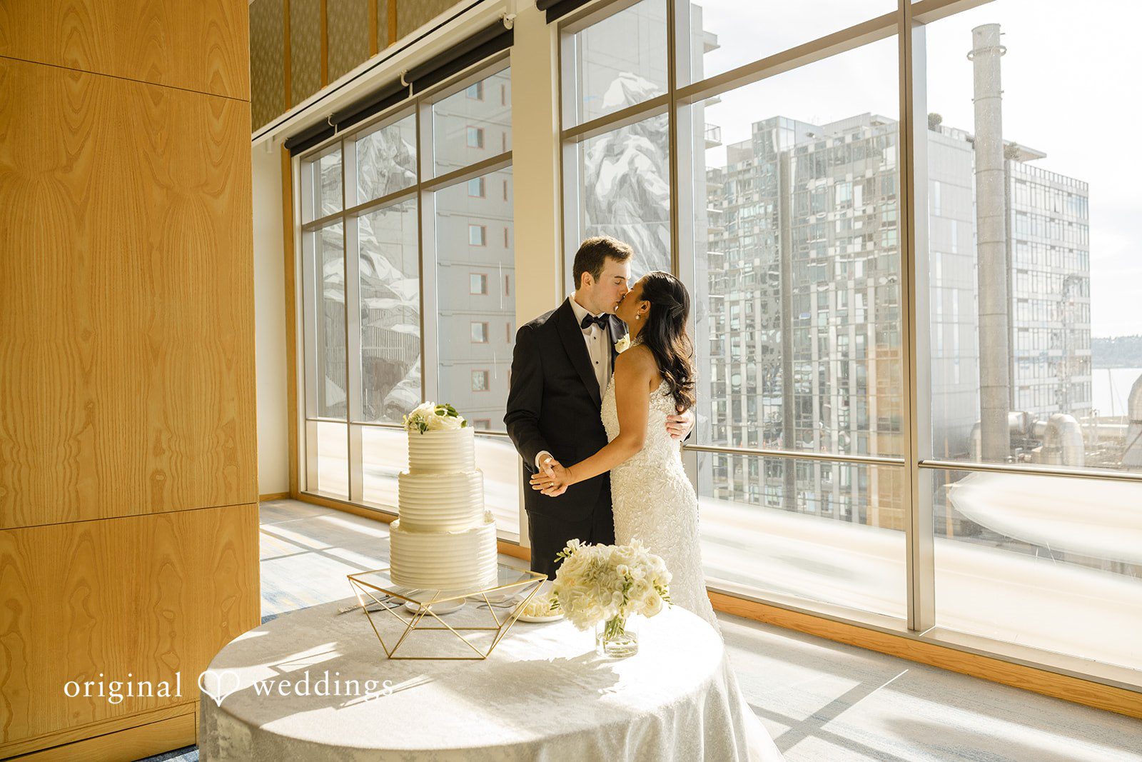 John + Michelle Bride and groom kissing next to a wedding cake display in front of tall windows.