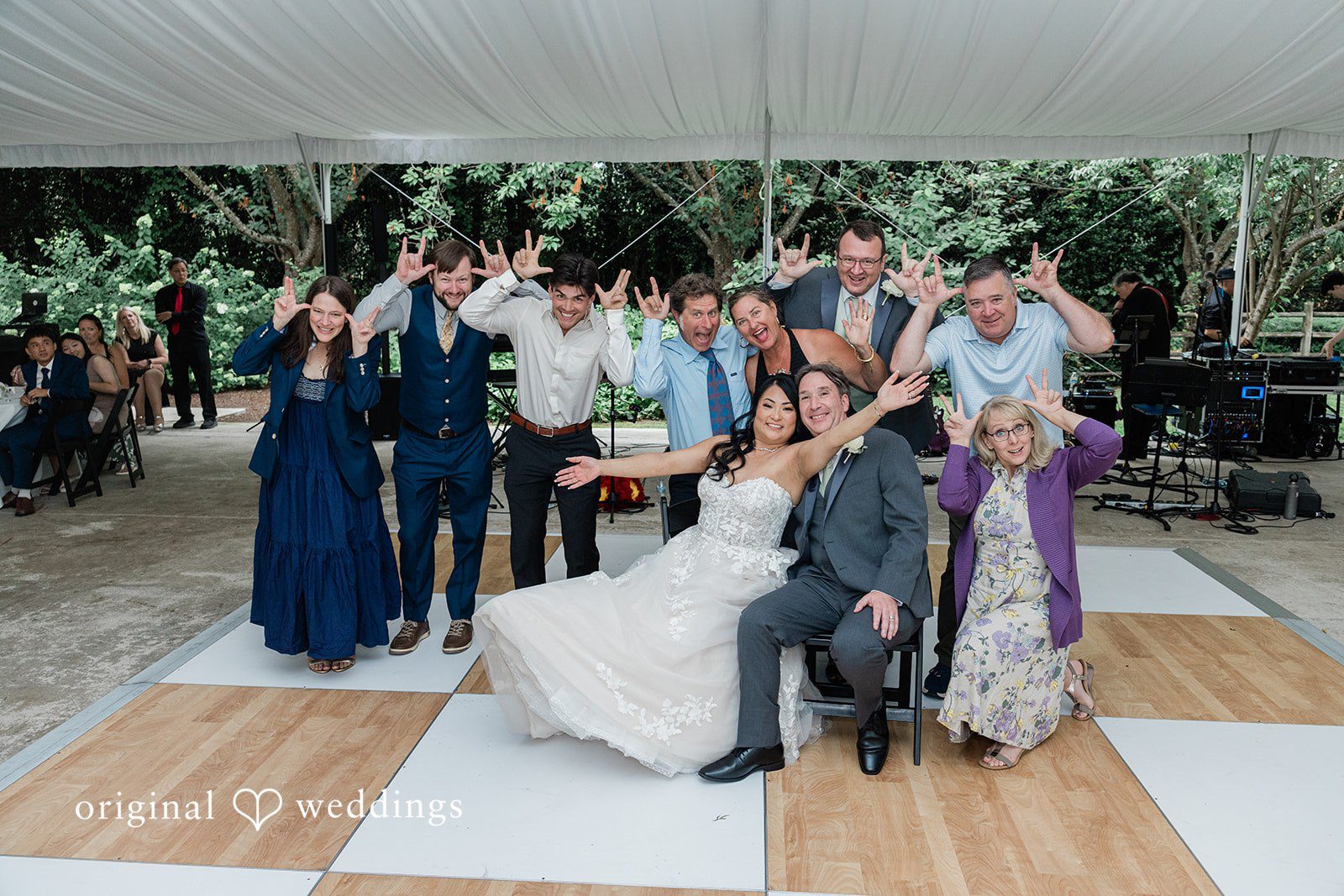 Kim + Keven A wedding party poses playfully on a checkered dance floor under a white tent, making hand gestures and smiling.