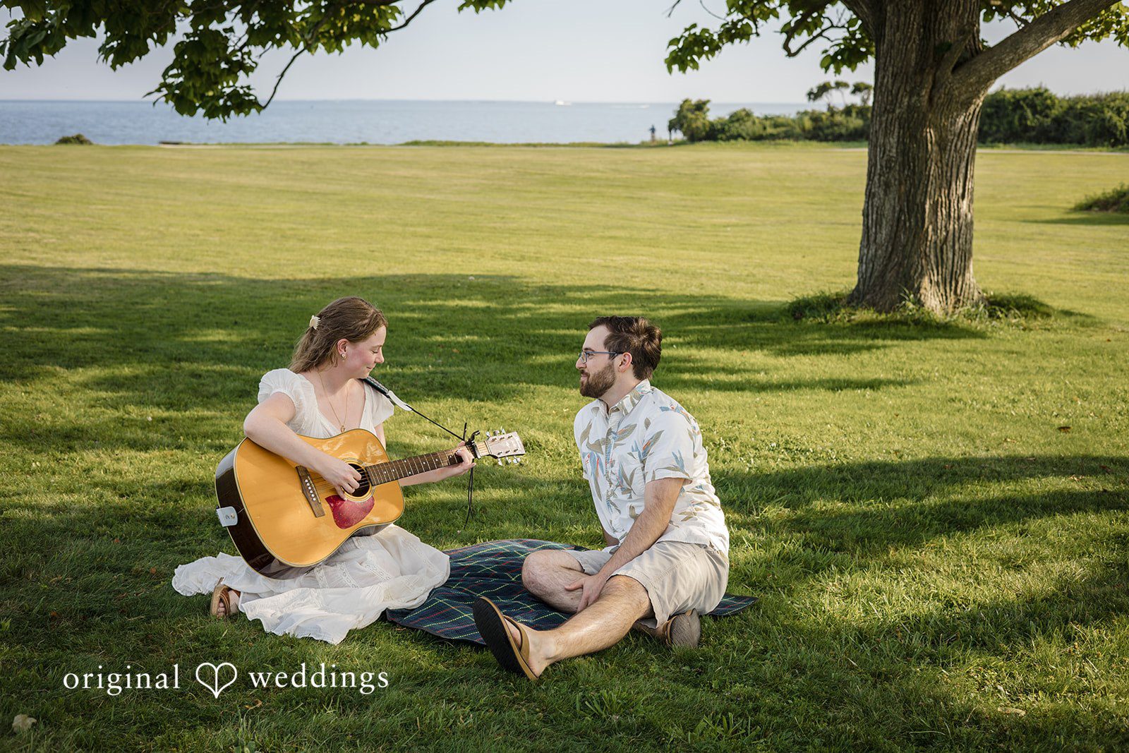 James + Ashlan A man sits on the grass while a woman sits opposite him playing an acoustic guitar.
