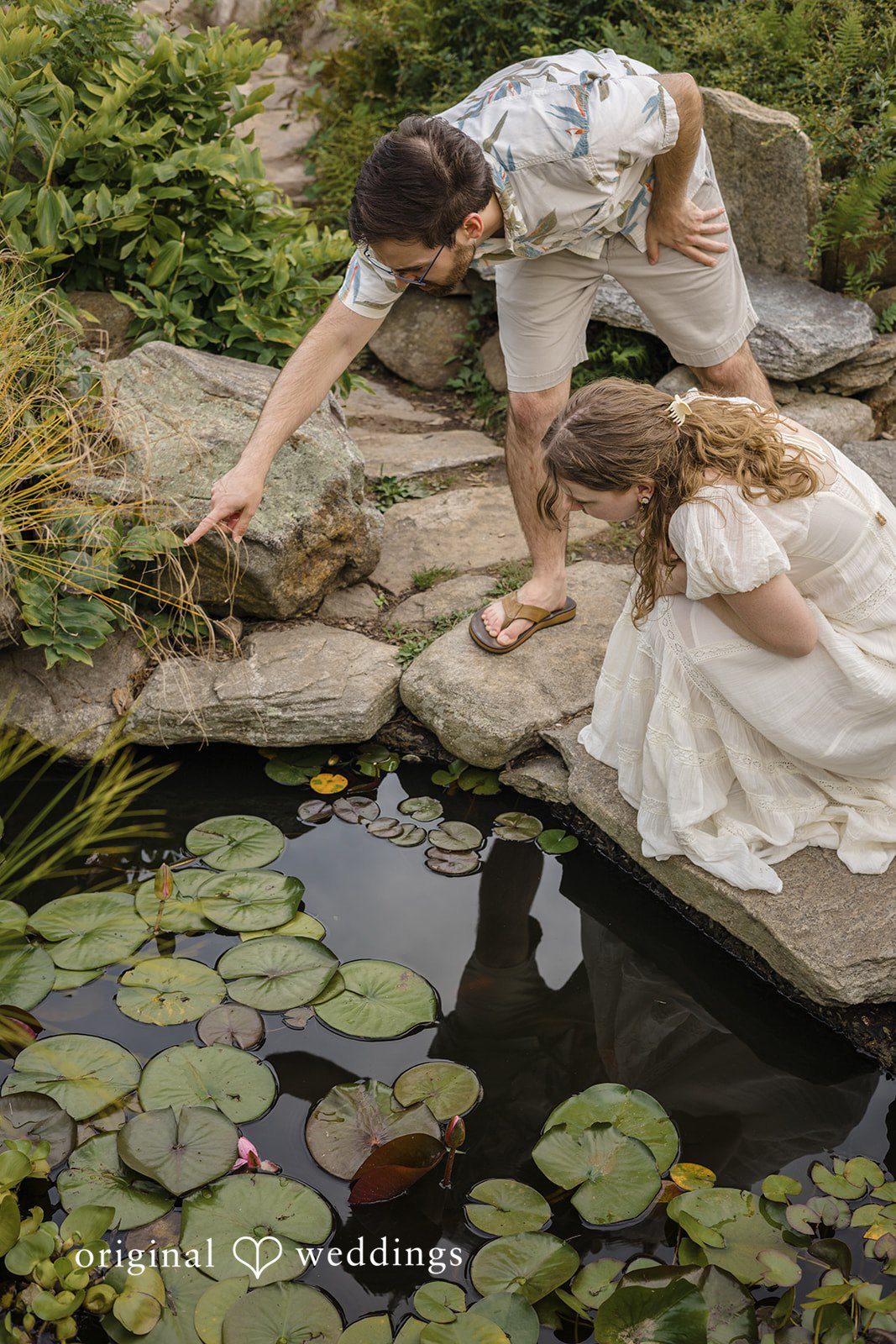 James + Ashlan A man leans down over a stone ledge toward a woman sitting by a rocky pond.