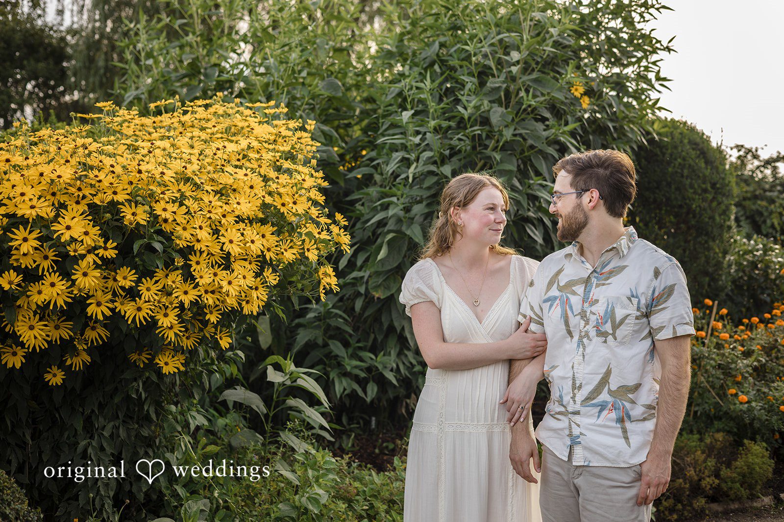 James + Ashlan A man and a woman stand side-by-side next to a large bush of yellow flowers.