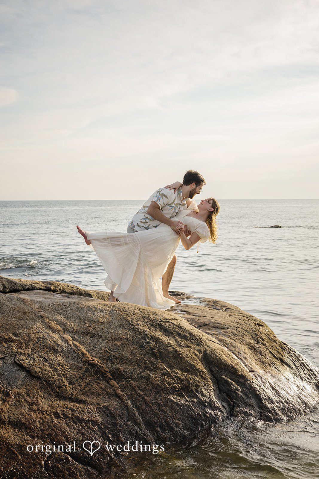 James + Ashlan A man lifts a woman up horizontally over large rocks near the water.