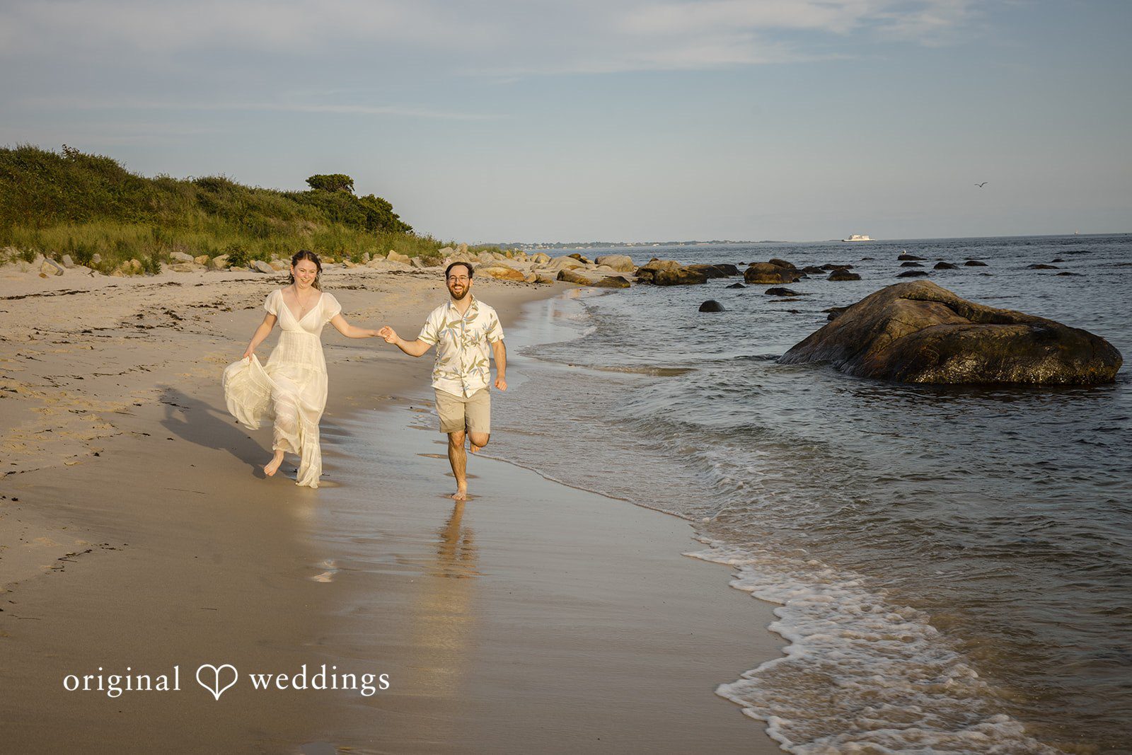 James + Ashlan Two people walk along a sandy shoreline next to the ocean.