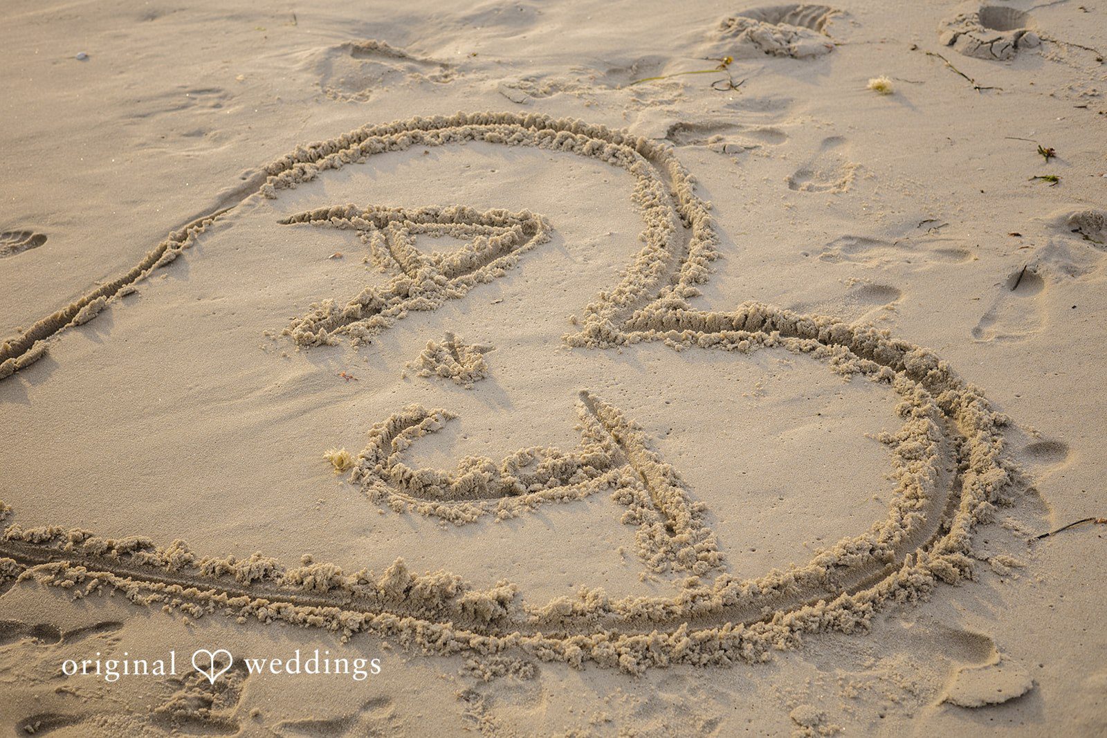 James + Ashlan A stylized heart symbol enclosing initials is drawn in the sand on a beach.