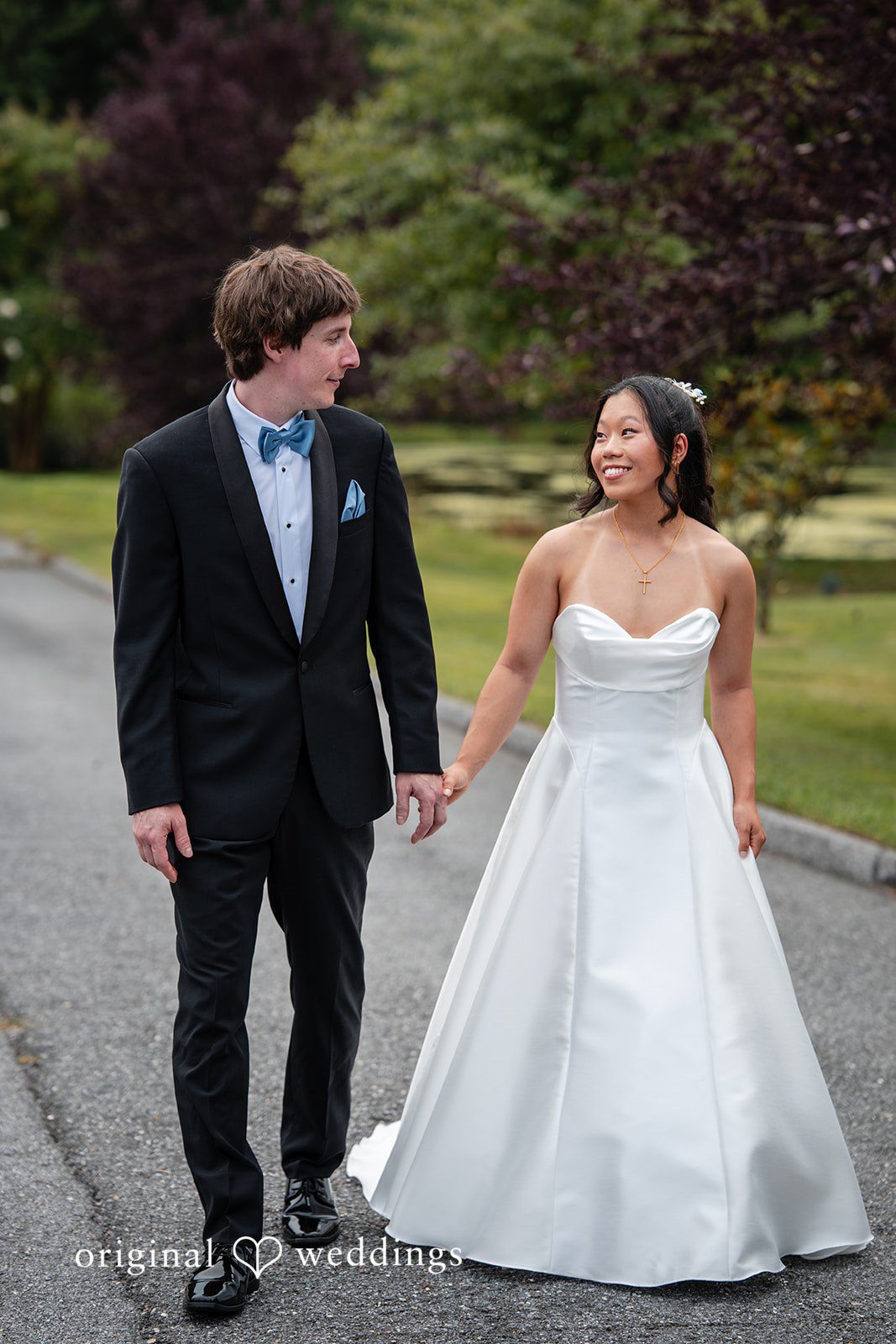 Mae + Corey A bride in a white gown and a groom in a black tuxedo walk hand-in-hand down a paved path.