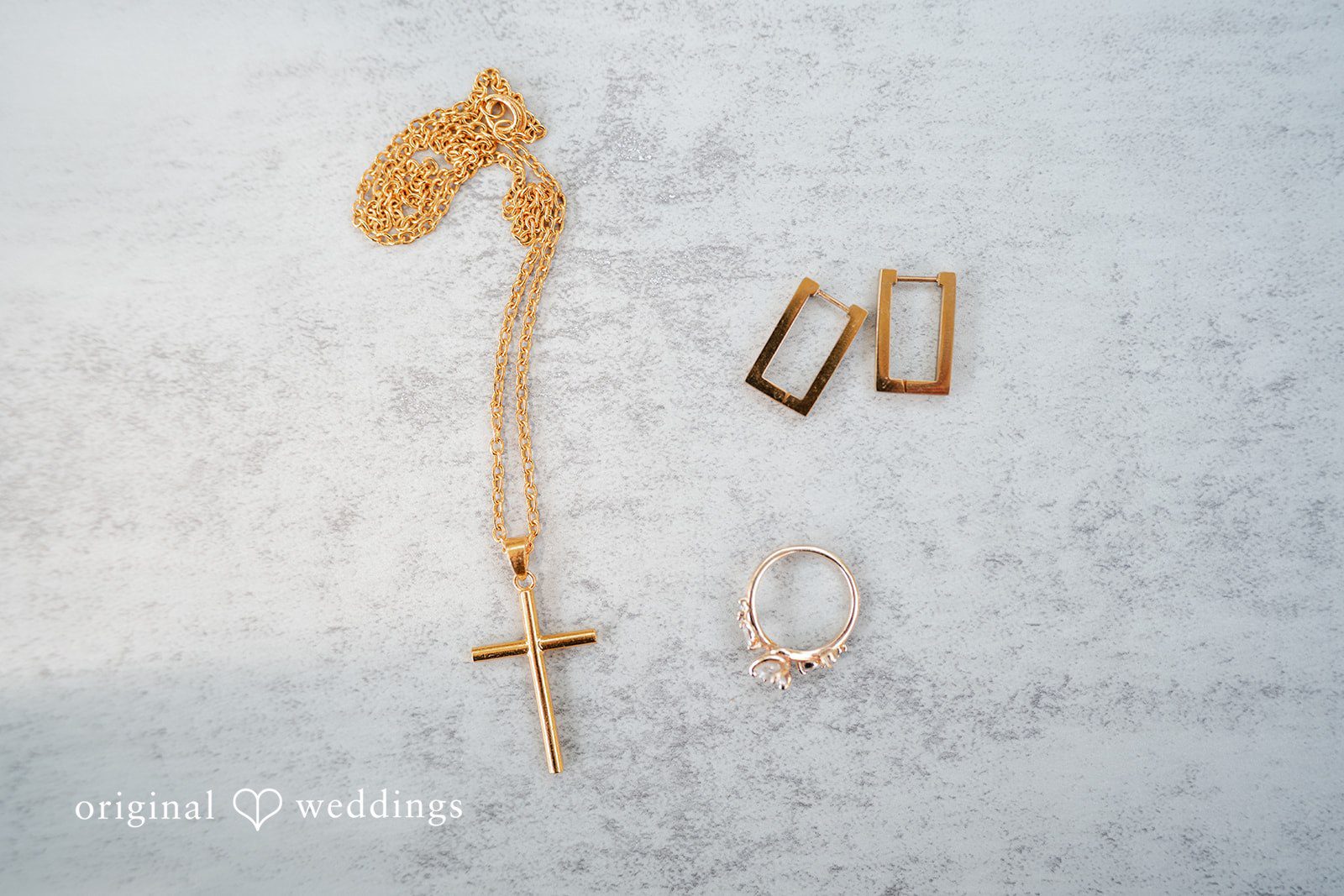 Mae + Corey A flat lay display of gold wedding jewelry and a cross pendant on a textured surface.