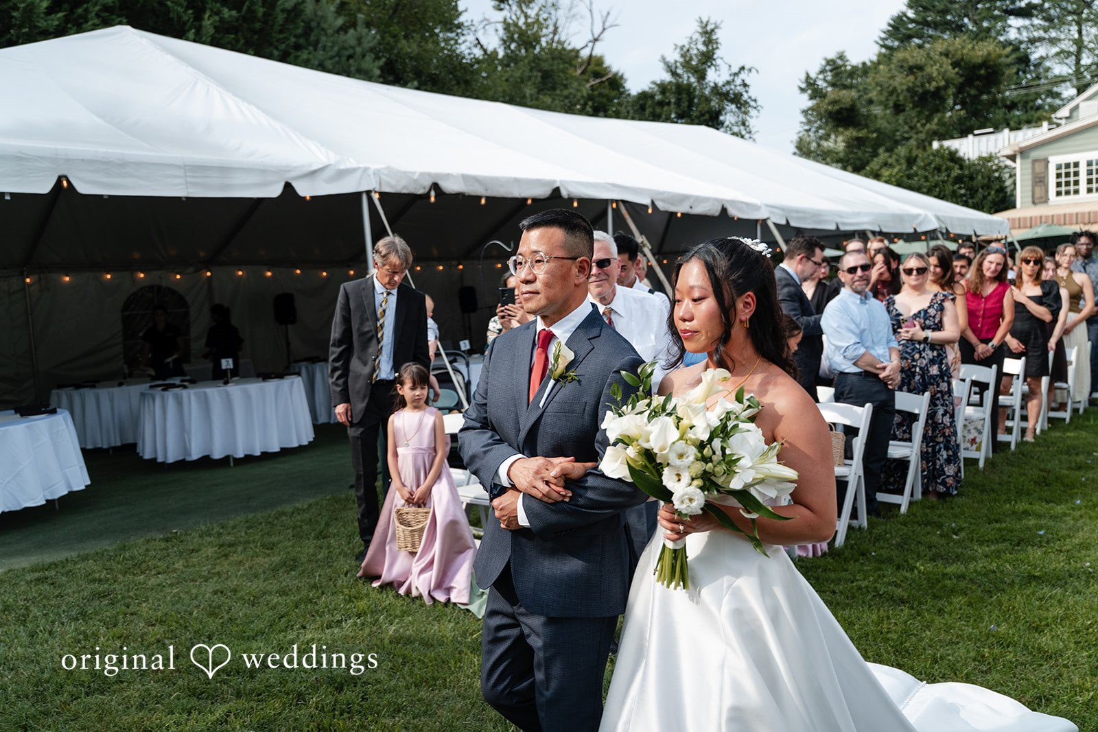 Mae + Corey A bride walks down the aisle arm-in-arm with a man as wedding guests watch.
