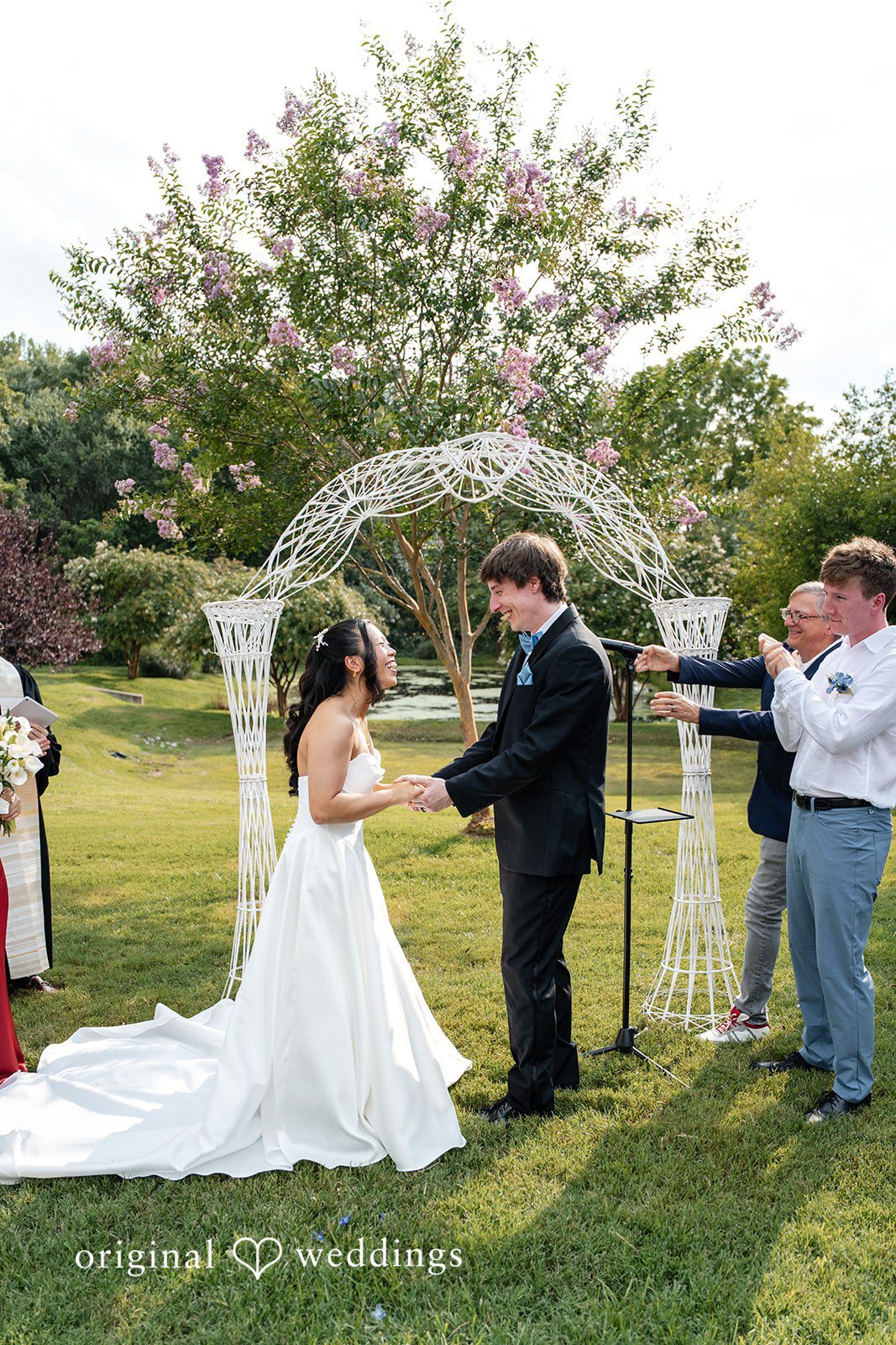 Mae + Corey A bride and groom hold hands while exchanging vows under a white trellis archway.