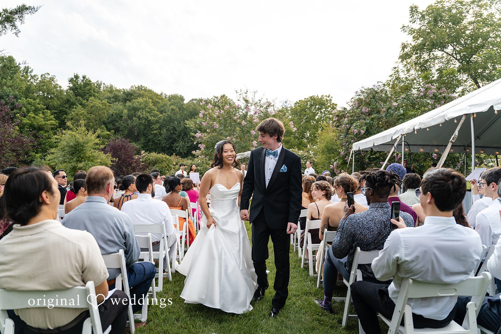Mae + Corey A bride and groom walk down the aisle together past cheering wedding guests.