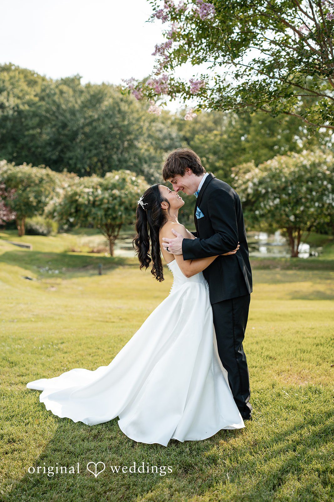 Mae + Corey A groom dips a bride backwards for a kiss on a green lawn surrounded by trees.
