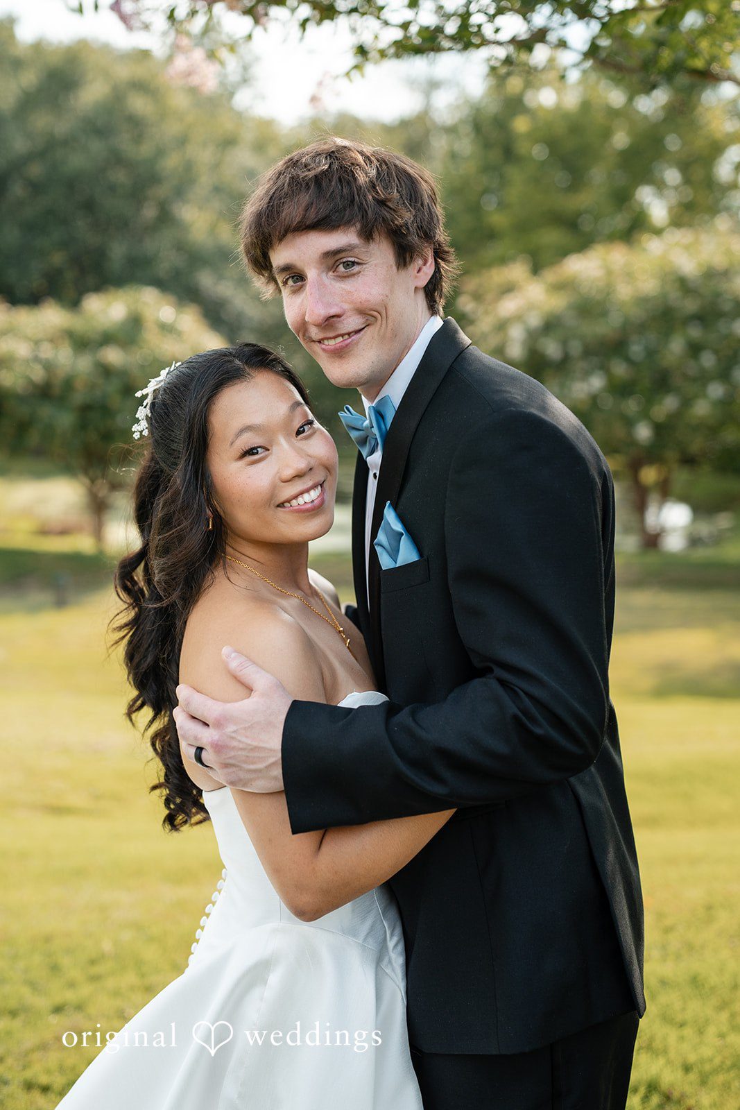 Mae + Corey A groom in a black tuxedo wraps his arms around a smiling bride from behind in a grassy field.