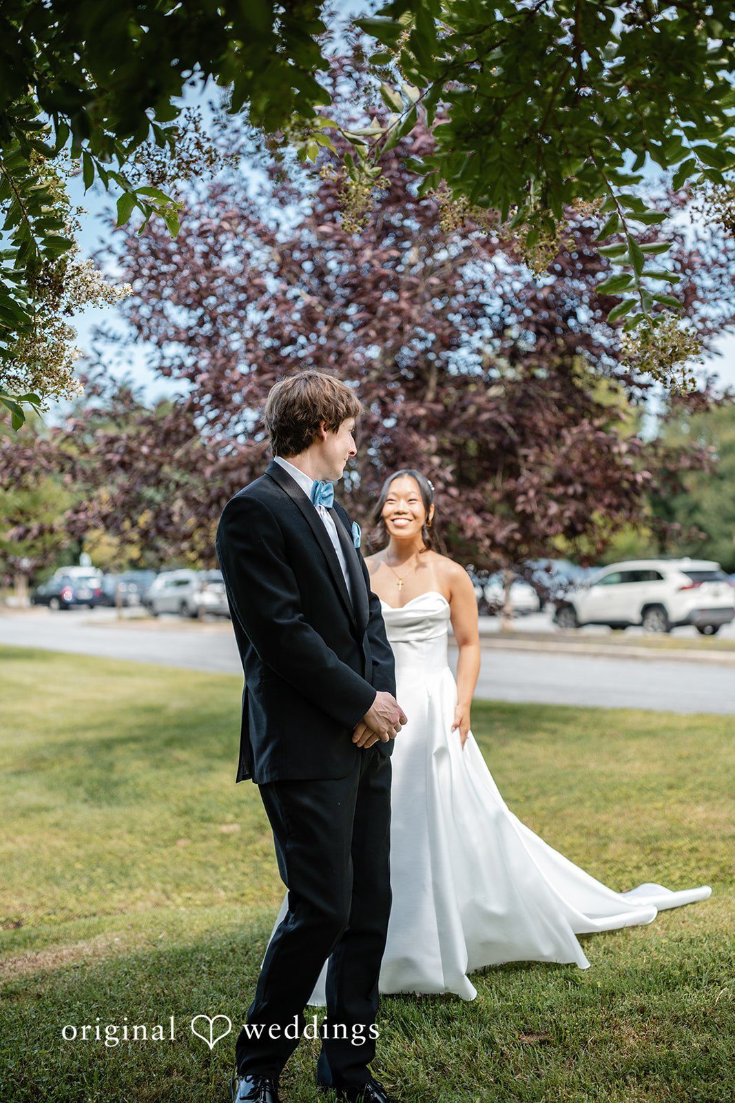 Mae + Corey A bride and groom pose together on a green lawn near a paved parking lot area.