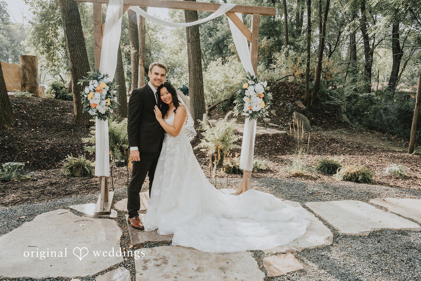 Jake + Ida Ida and Jake posing under the floral ceremony arch, captured by Minneapolis wedding photographers from Original Weddings at Lake Vadnais Grand Events