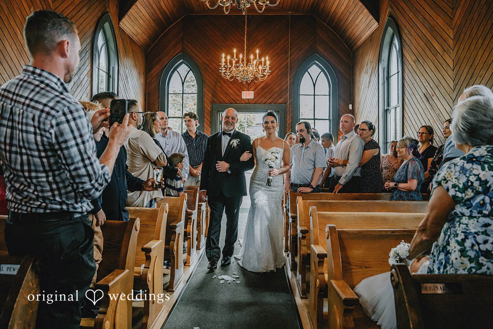 Ashley + Forrest A bride walks down a church aisle past wedding guests seated in wooden pews.