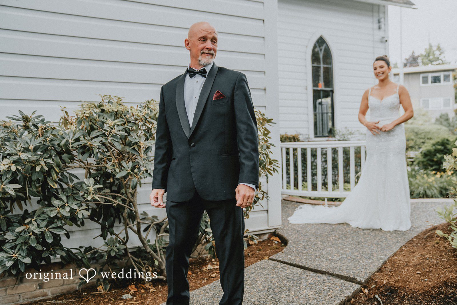 Ashley + Forrest A man in a dark suit stands on a walkway outside a church while a woman in a white dress stands in the background.