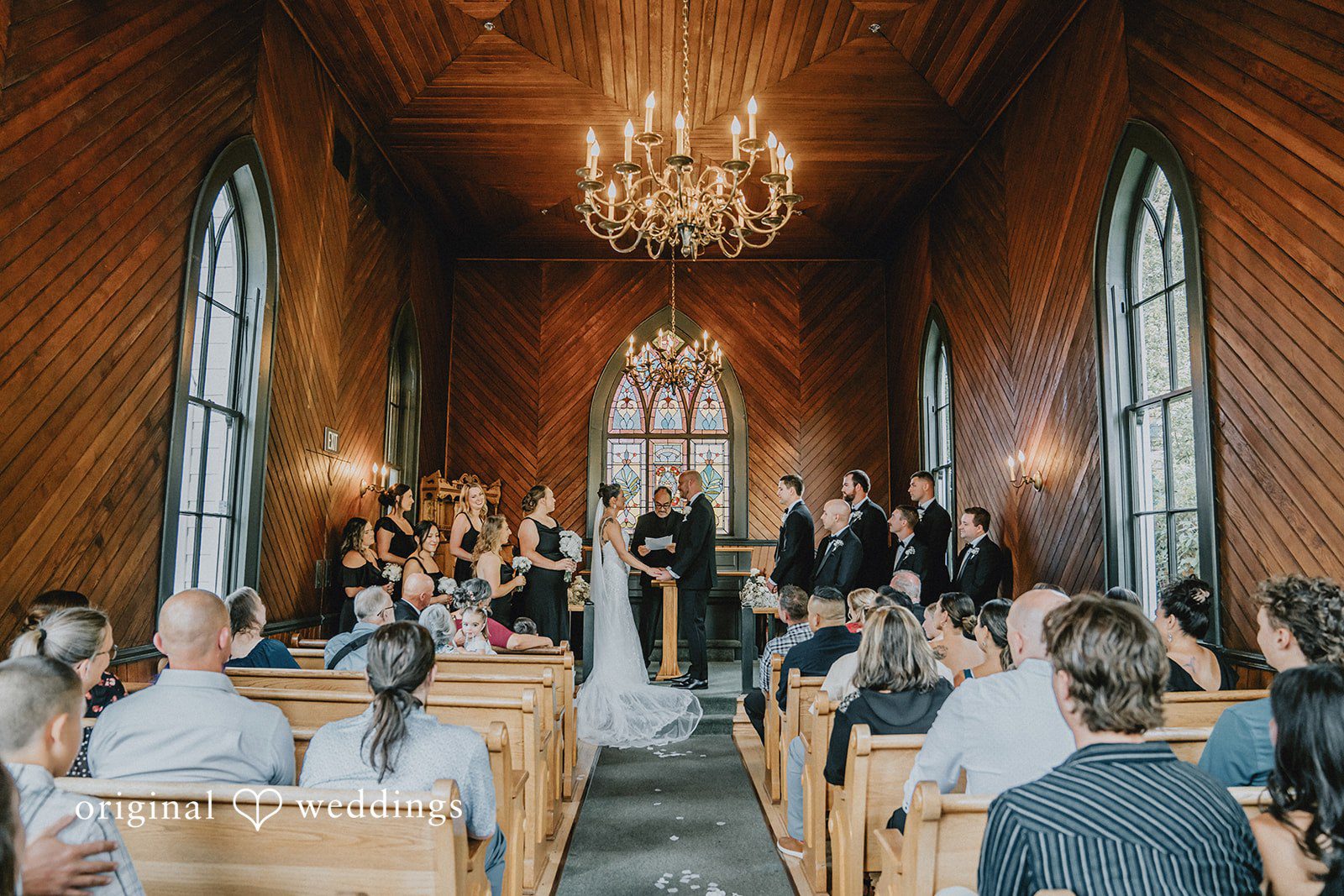 Ashley + Forrest A wide view of a wedding ceremony inside a wood-paneled church filled with seated guests.