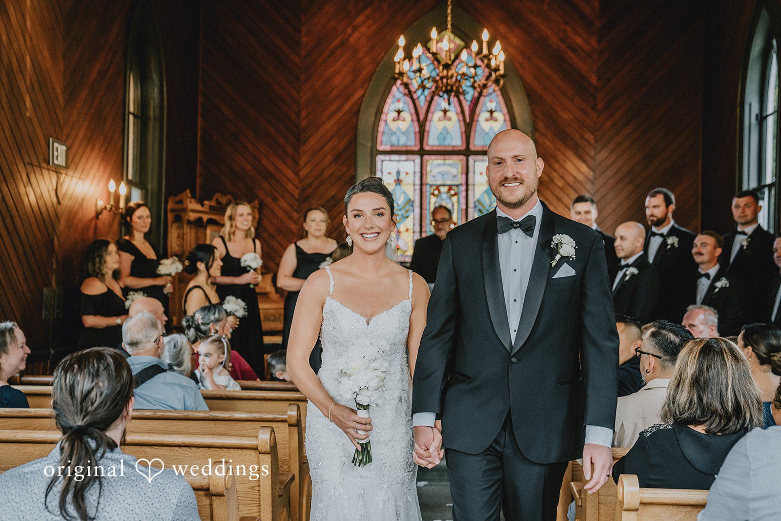 Ashley + Forrest A bride and groom stand side-by-side smiling at their wedding guests inside a church.