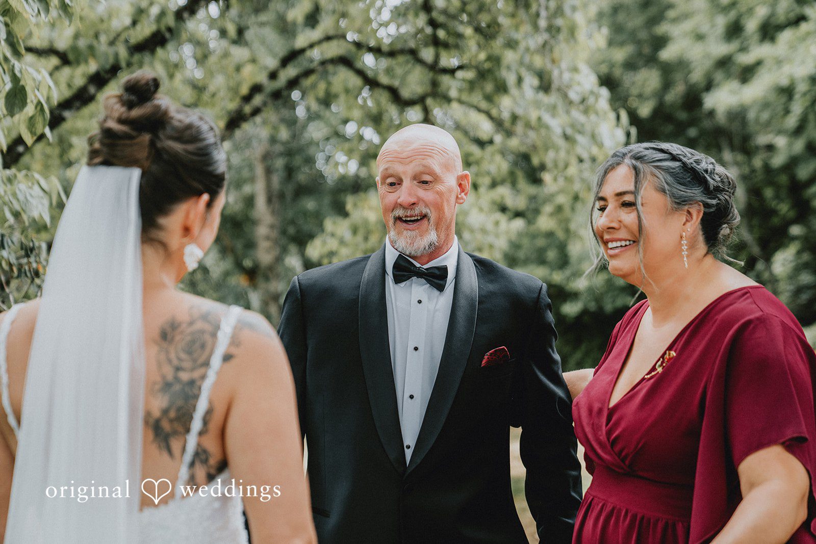 Ashley + Forrest A man in a black tuxedo smiles while standing between two women at a wedding venue