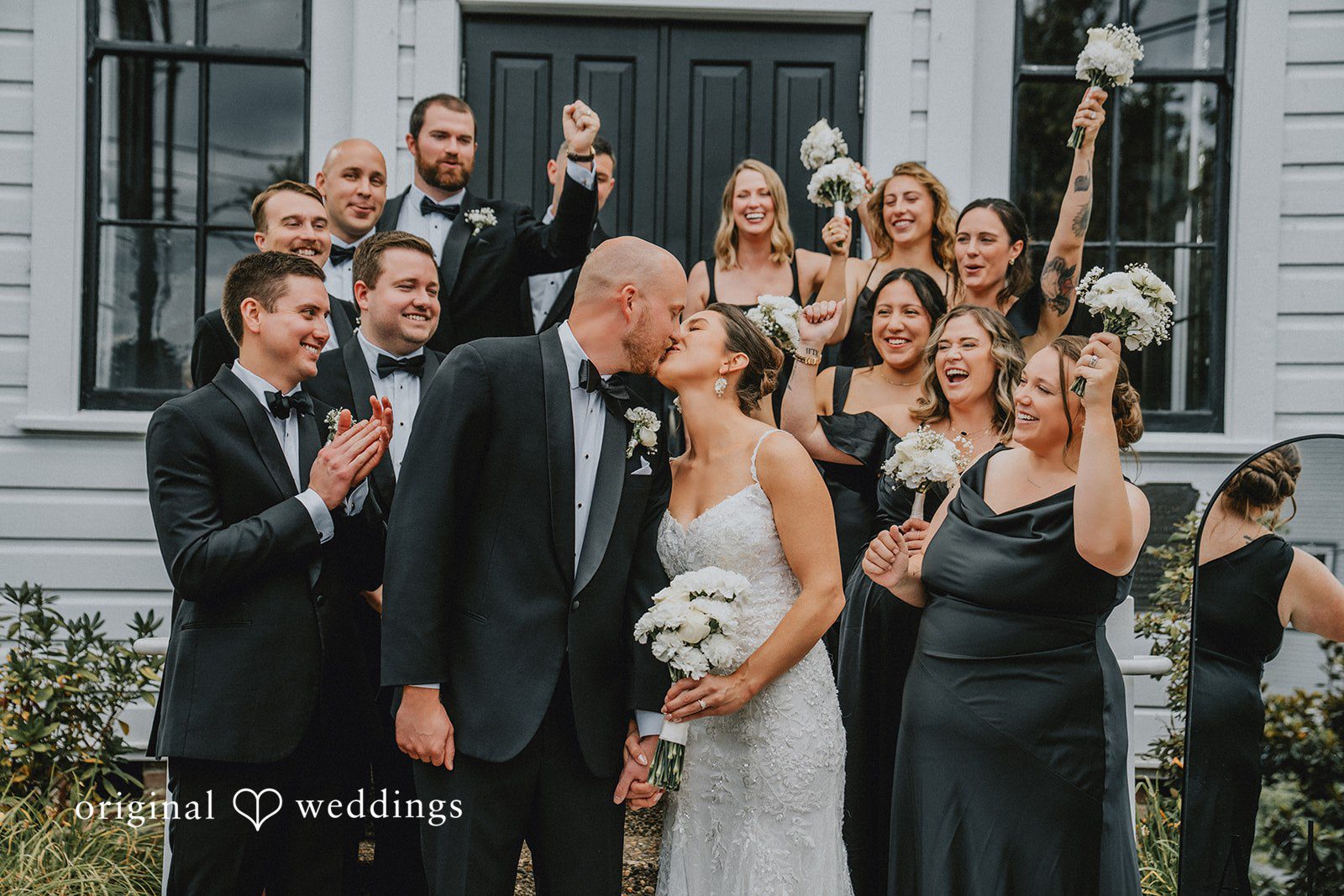 Ashley + Forrest A bride and groom kiss while surrounded by cheering guests on an outdoor walkway.