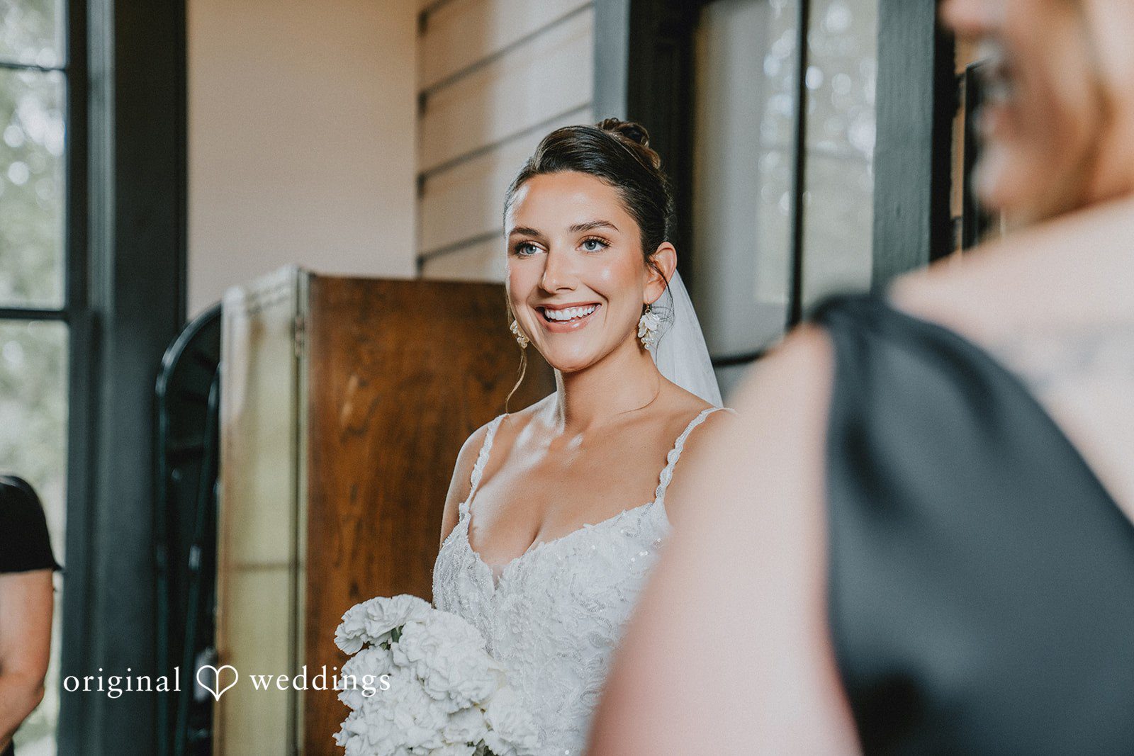 Ashley + Forrest A smiling bride in a white gown is seen over the shoulder of another person.
