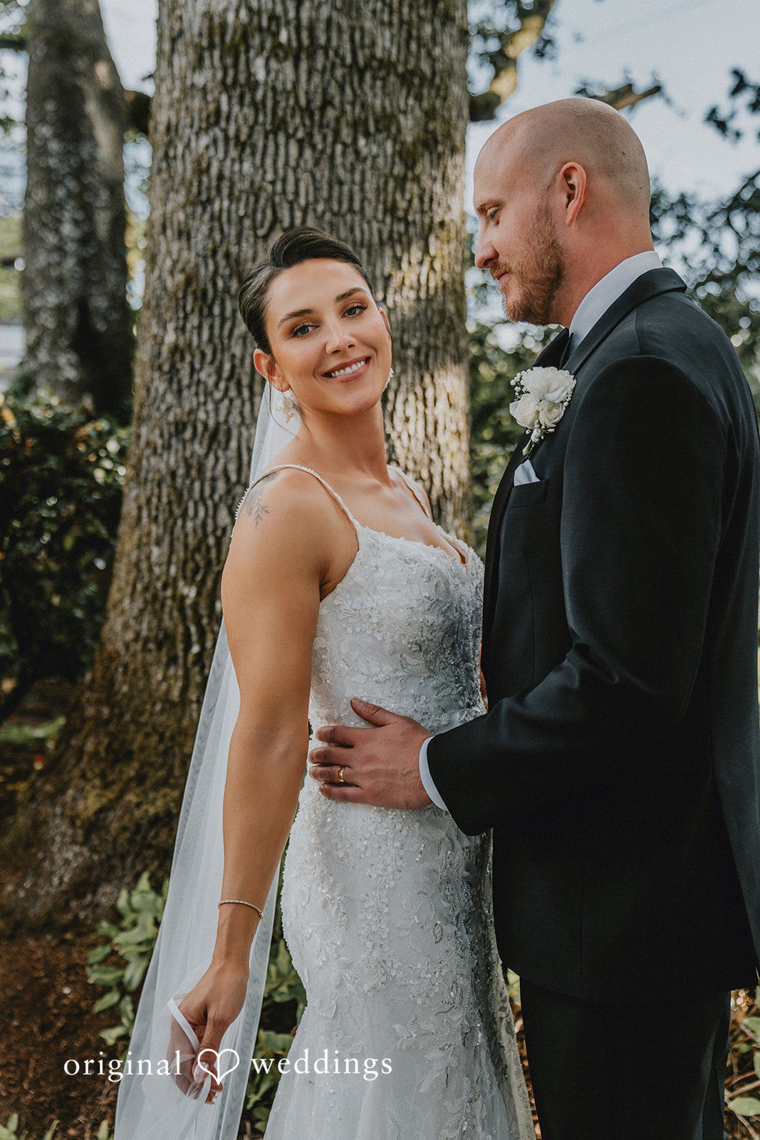 Ashley + Forrest A bride in a white gown and a groom in a black tuxedo smile at each other while posing.