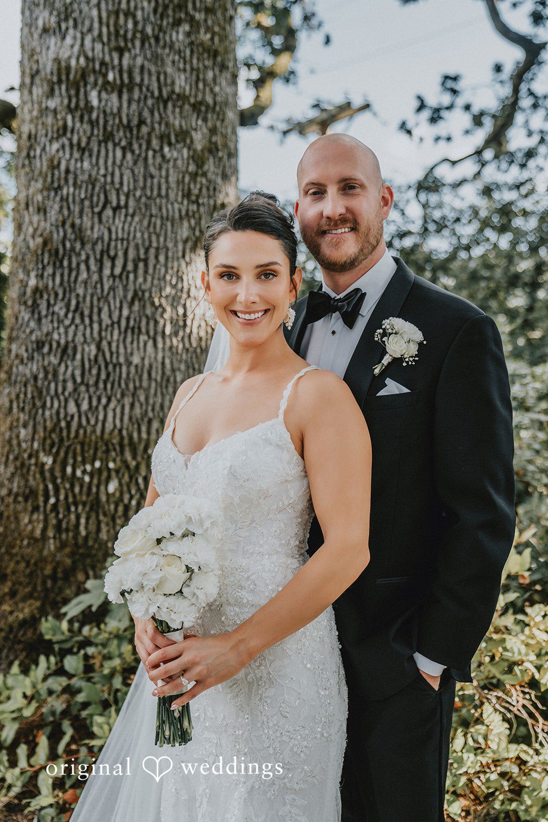 Ashley + Forrest A bride and groom stand close together smiling for a portrait against a backdrop of trees.
