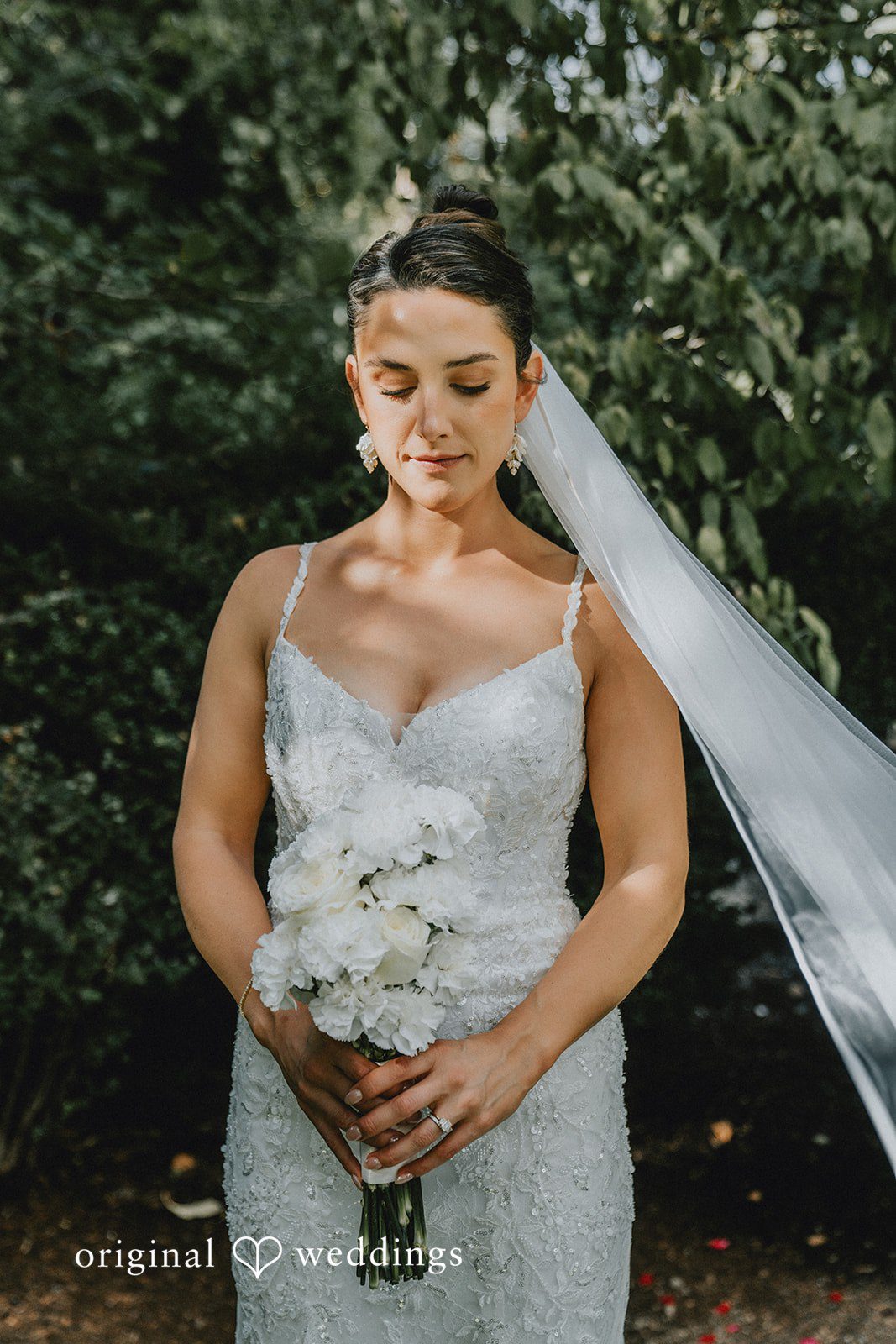 Ashley + Forrest A smiling woman in a textured white wedding gown looks downward while a veil drifts around her.