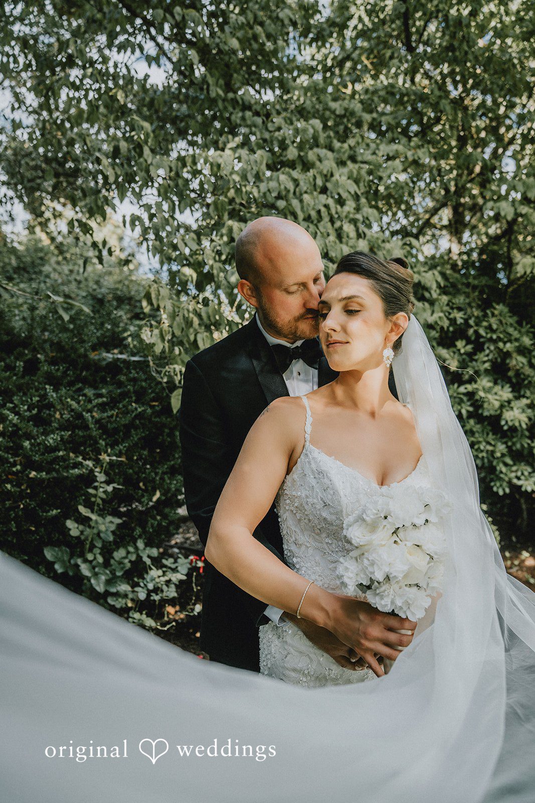 Ashley + Forrest A groom wraps his arms around a smiling bride from behind in front of dense green trees.