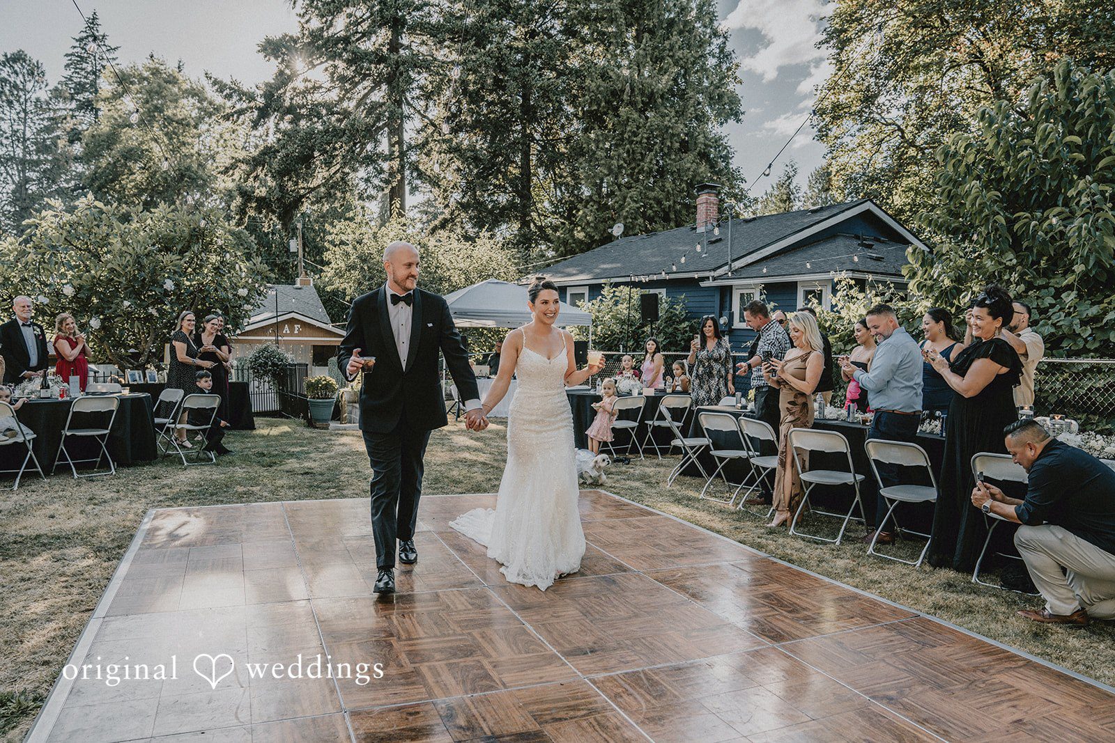 Ashley + Forrest A bride and groom hold hands while walking together across an open-air wooden dance floor.