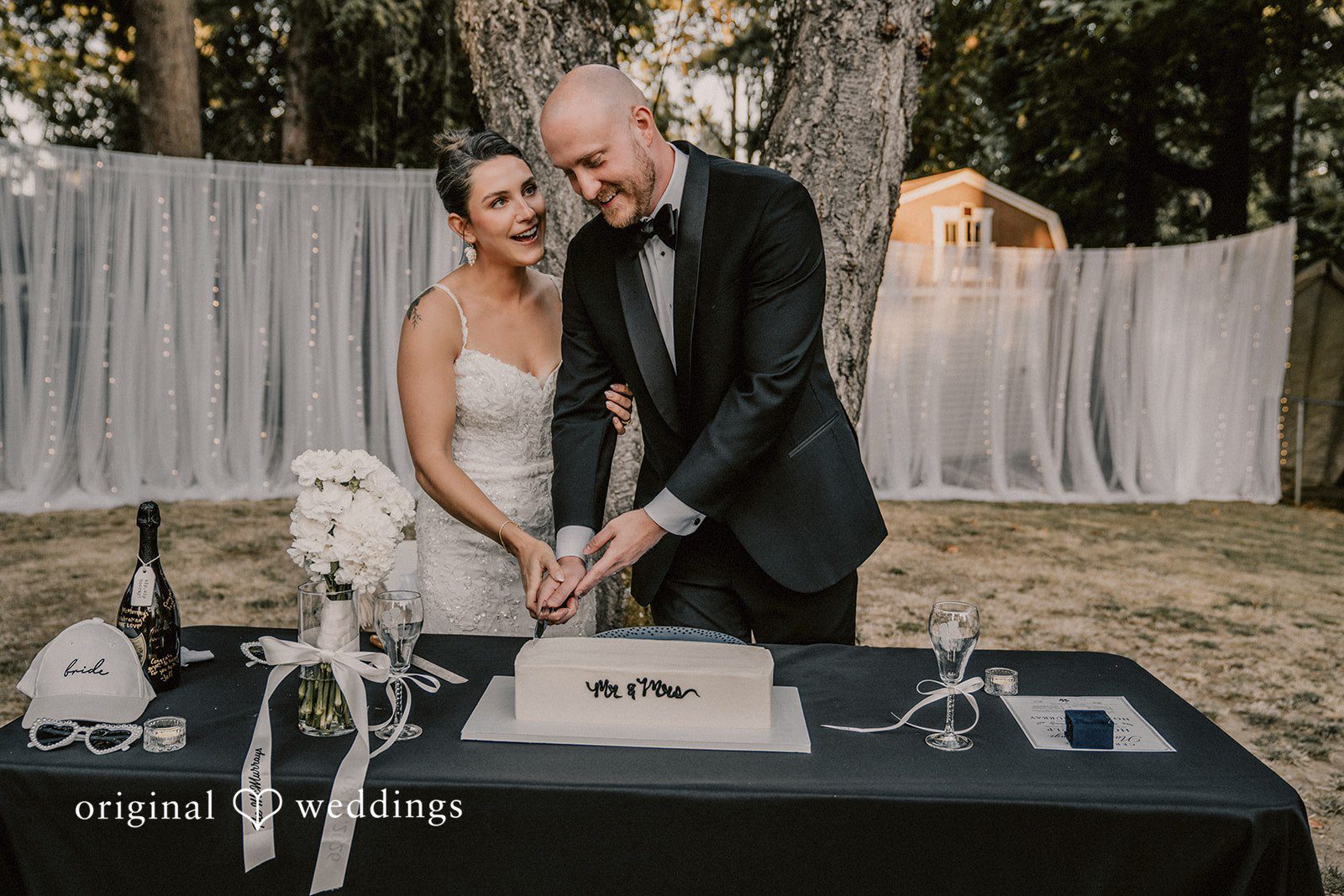 Ashley + Forrest A bride and groom cut a decorated white wedding cake together at a reception table.