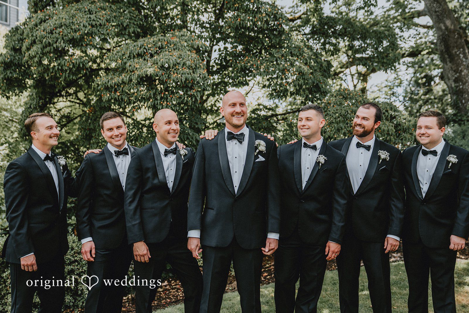 Ashley + Forrest A lineup of groomsmen in black tuxedos stands side-by-side in front of green trees.