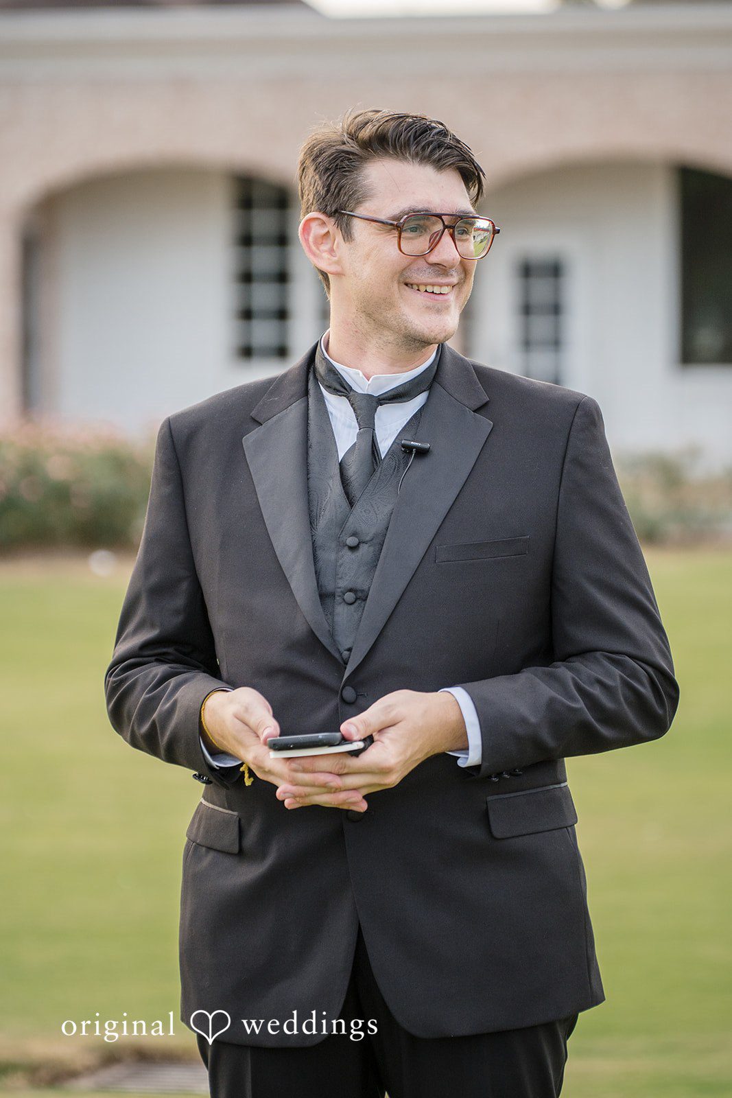 Nicholas + Nisha Groom in a black suit standing on a lawn and smiling.