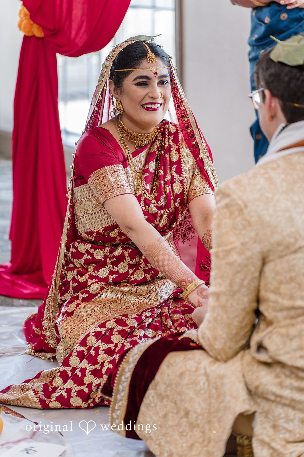 Nicholas + Nisha Two people sitting in a traditional wedding ritual under a red canopy.