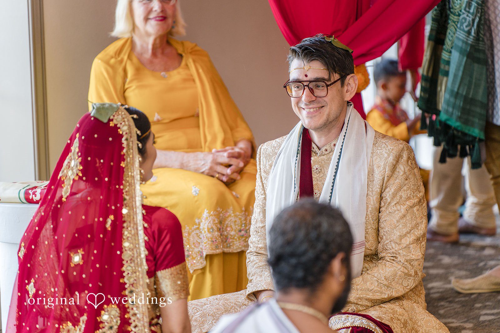 Nicholas + Nisha Groom smiling during a ceremony while seated among guests.