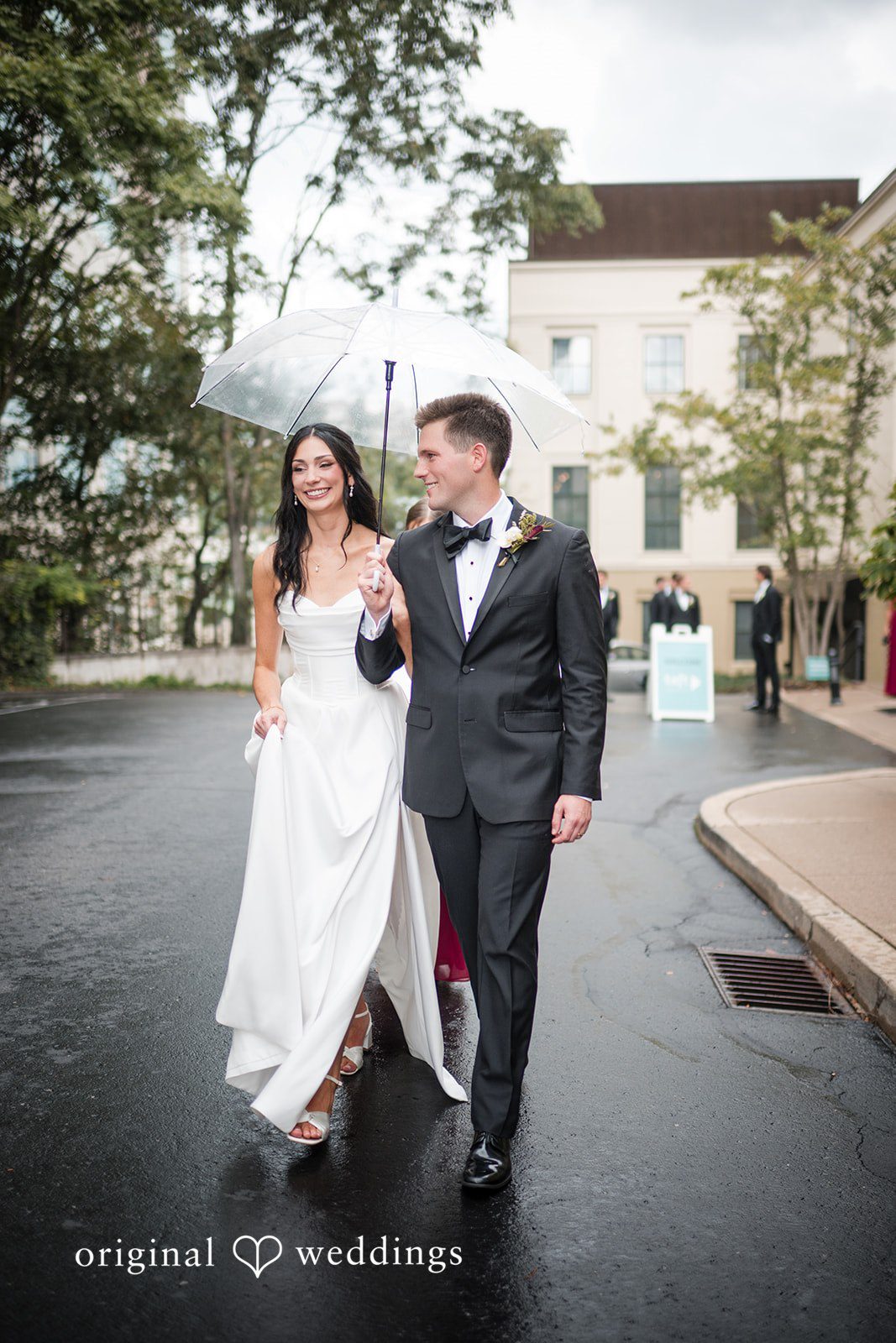 Macy + Nolan A bride and groom walking together along a paved path next to a building.