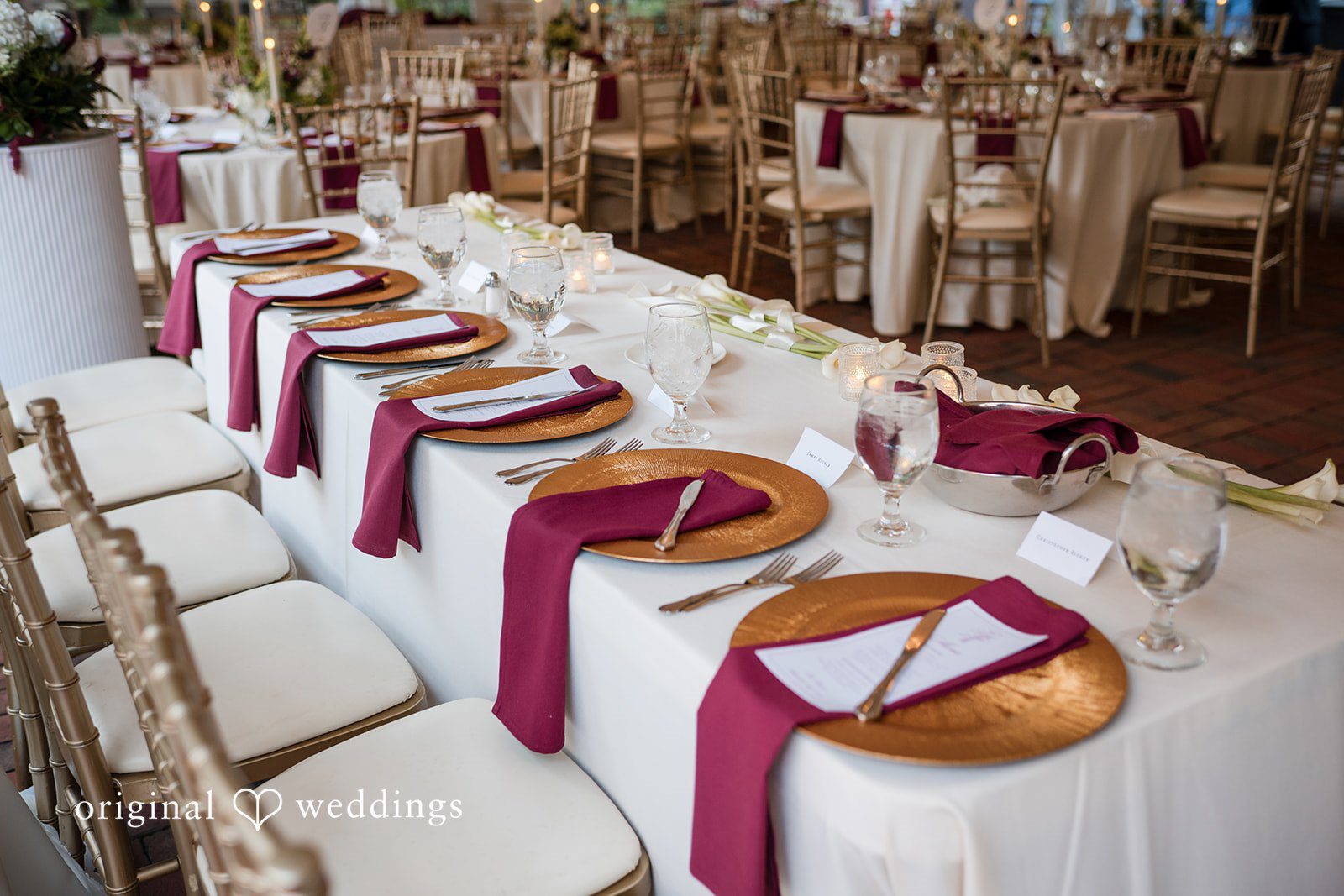 Macy + Nolan A long banquette-style reception table set with gold chargers and burgundy napkins.
