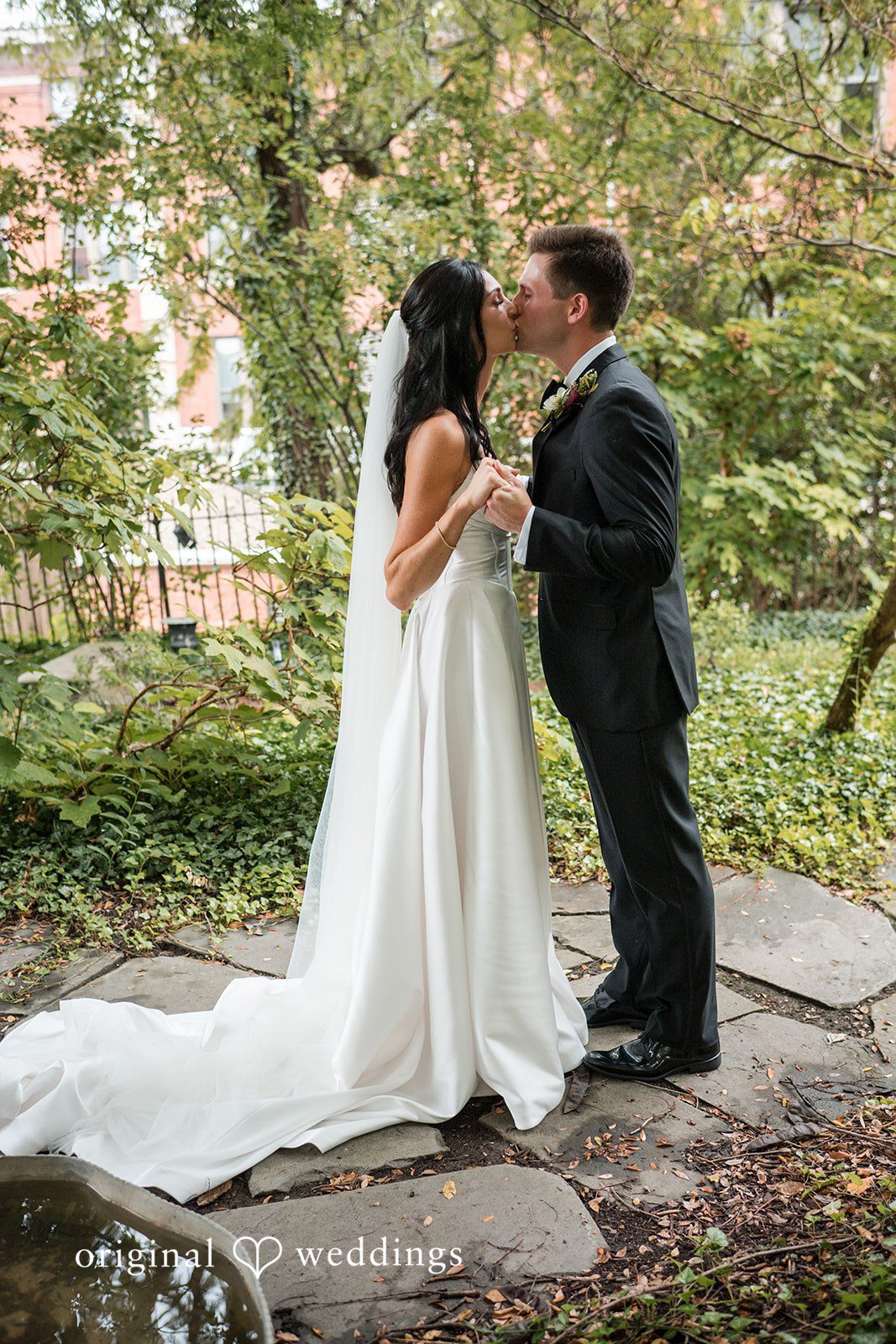 Macy + Nolan A bride and groom embracing on a grassy lawn next to trees.